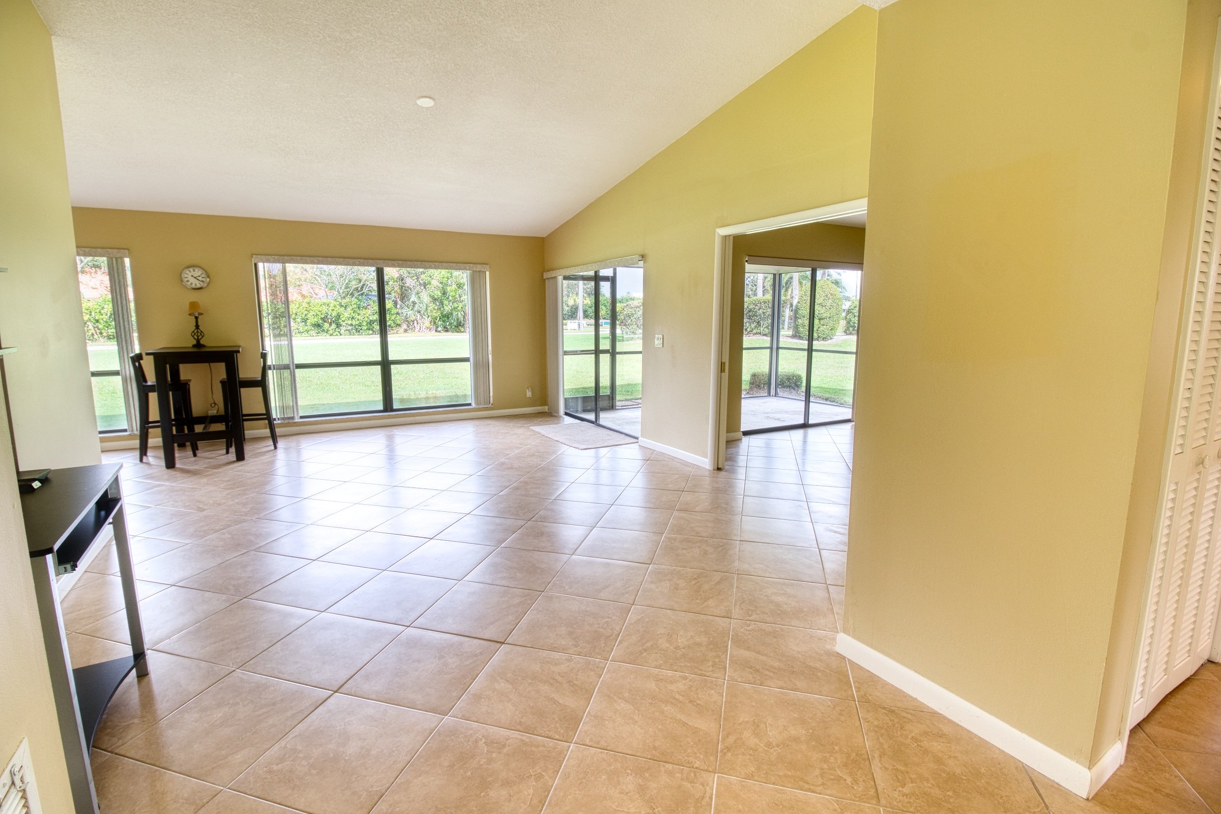 Empty living room with beige tiled floor, yellow walls, large windows, and sliding glass doors opening to a green lawn.