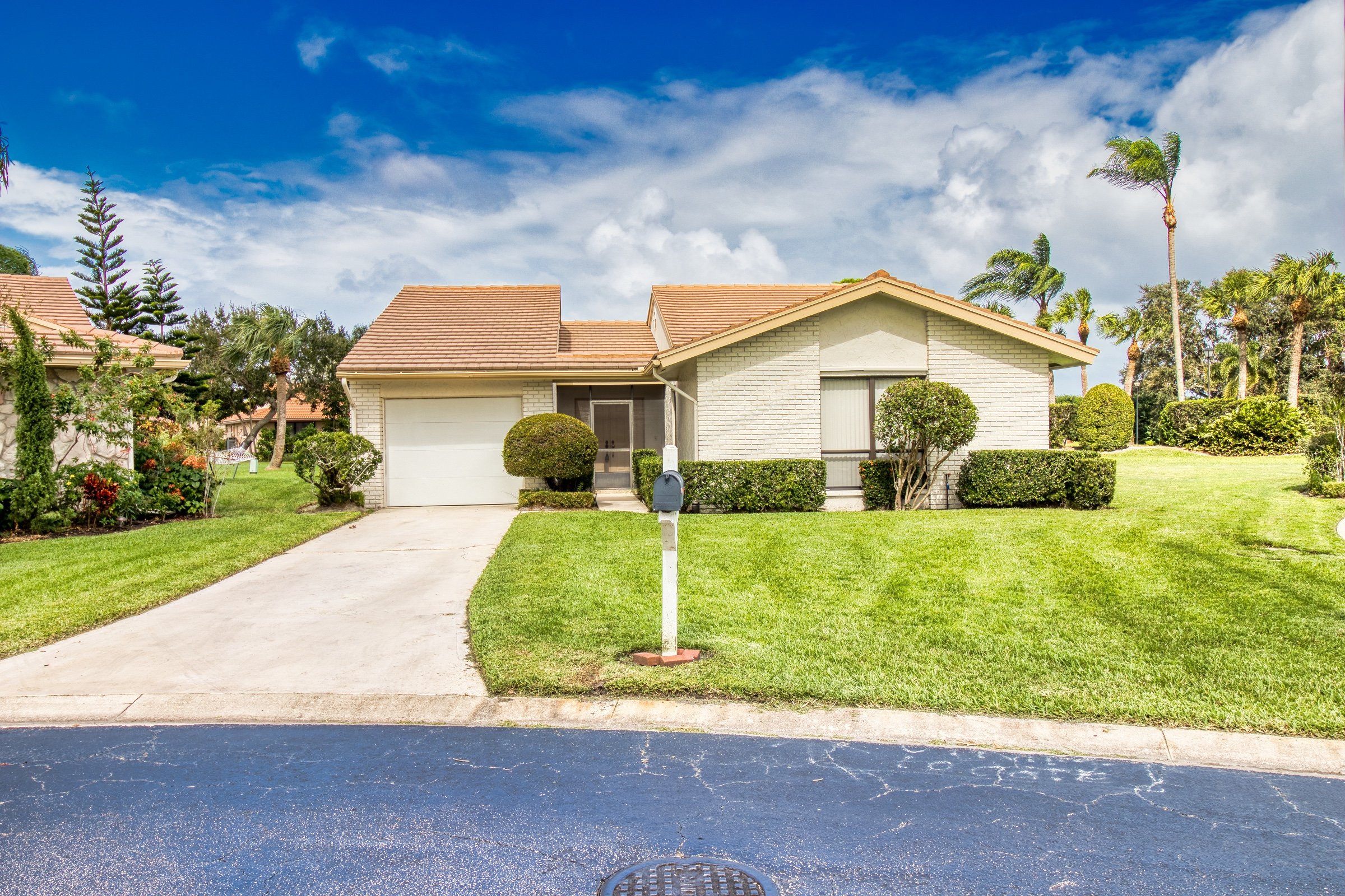 Suburban house with garage, manicured lawn, and mailbox, surrounded by palm trees under a blue sky.