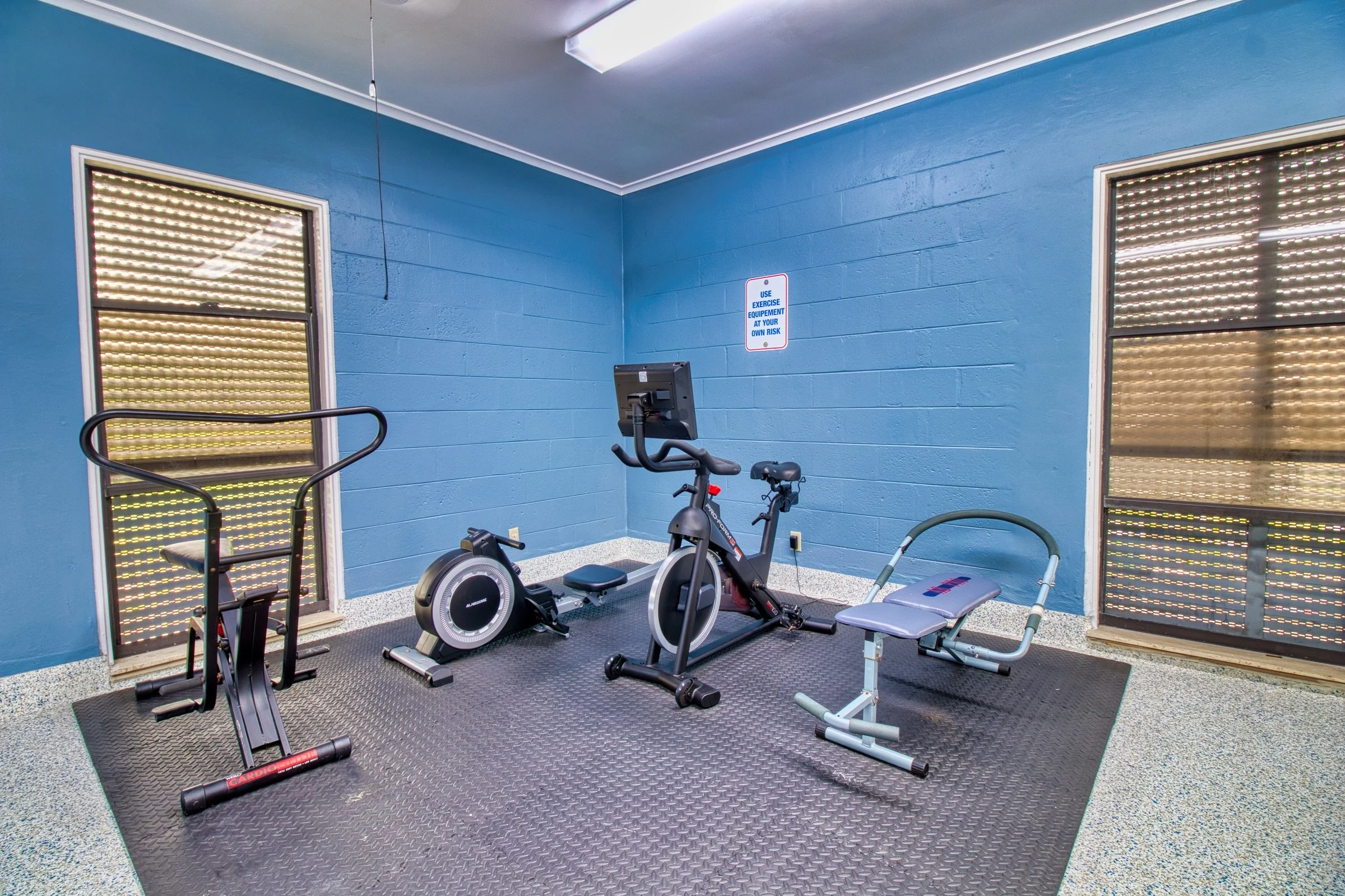 Small gym room with blue walls, featuring exercise equipment including a stationary bike, rowing machine, and ab bench.