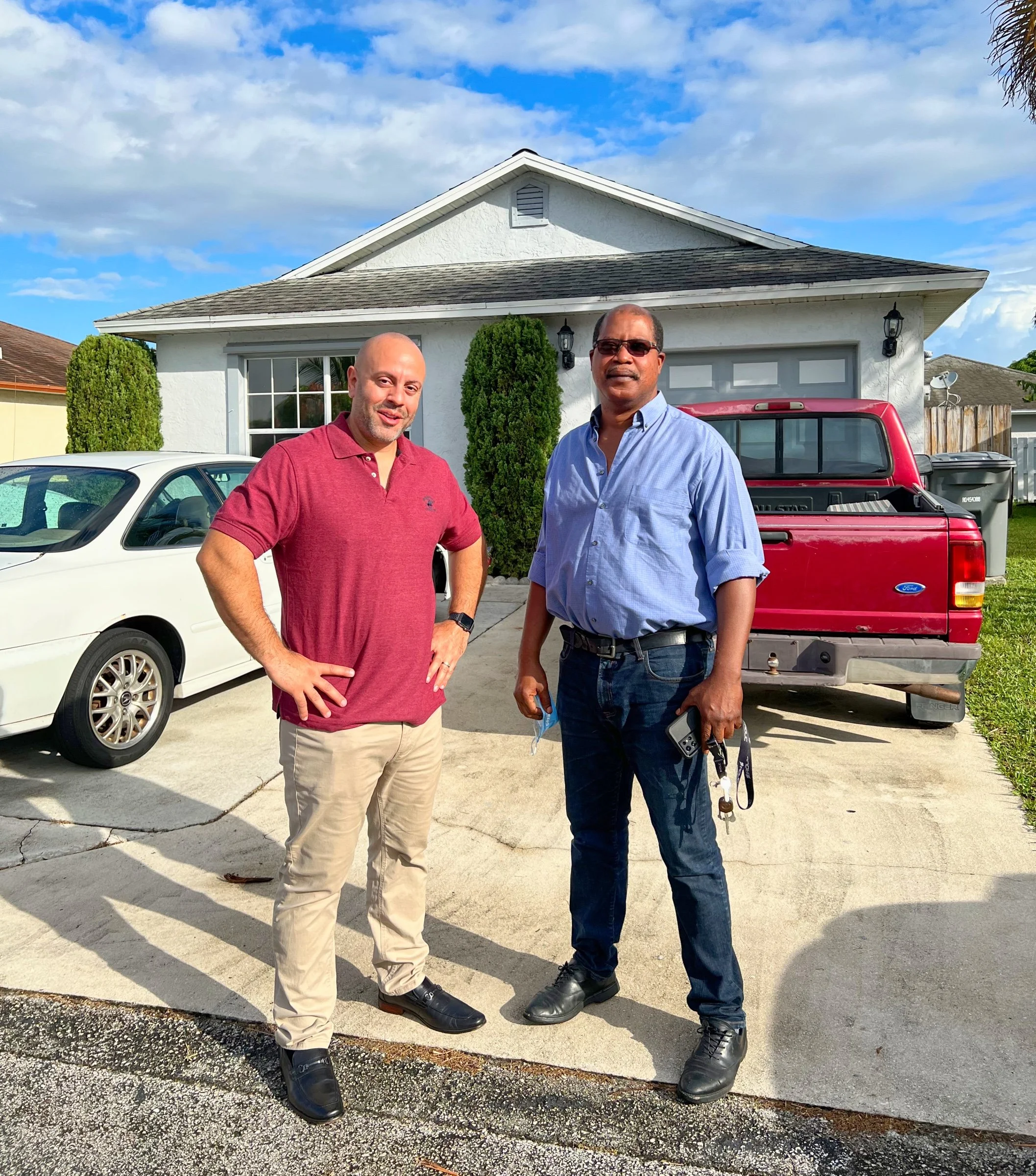 Two men standing on a driveway in front of a house with a white car and red truck.