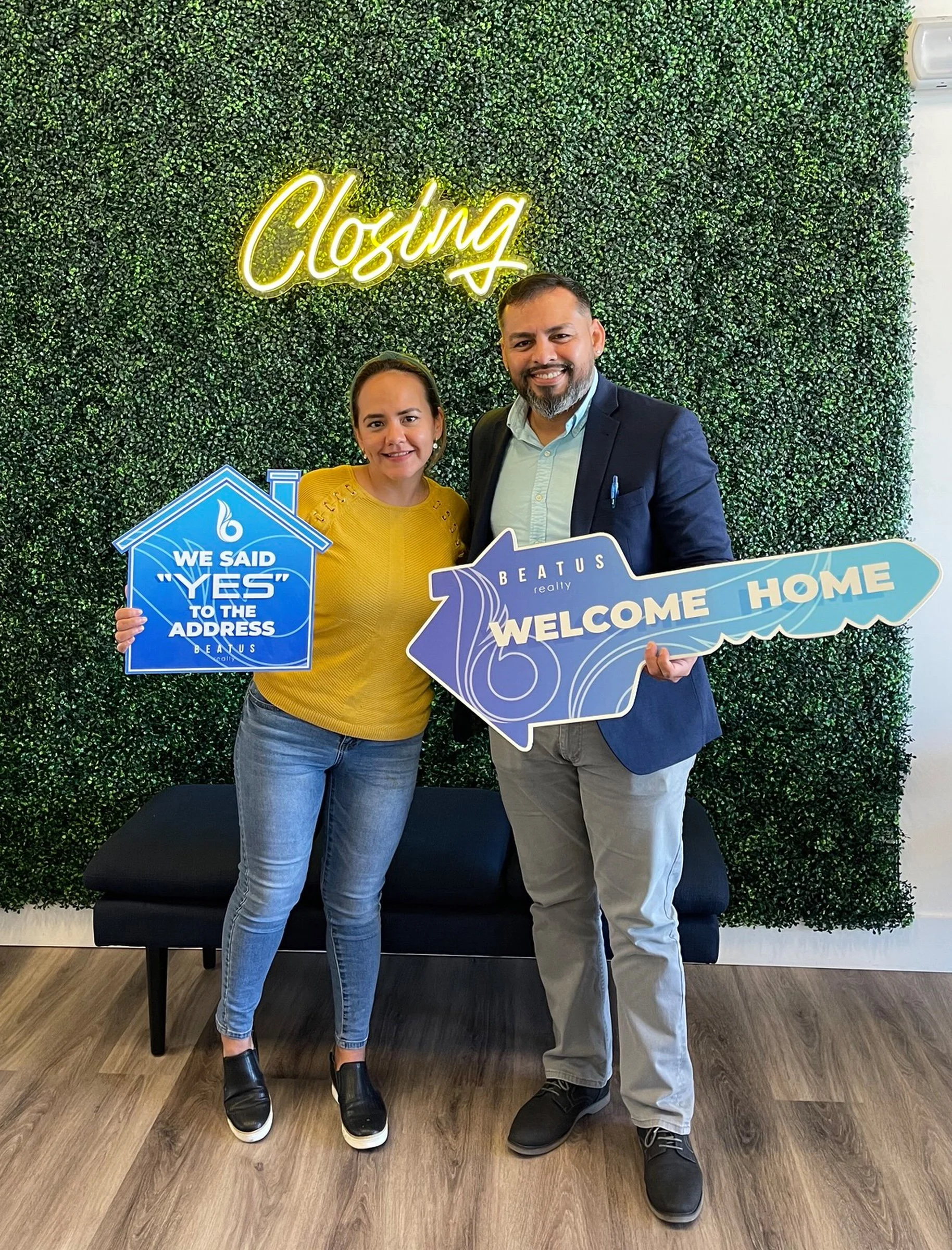 Two people holding signs in front of a green wall with a "Closing" neon sign. One sign says "We said yes to the address" and the other is a large key-shaped sign reading "Welcome Home."