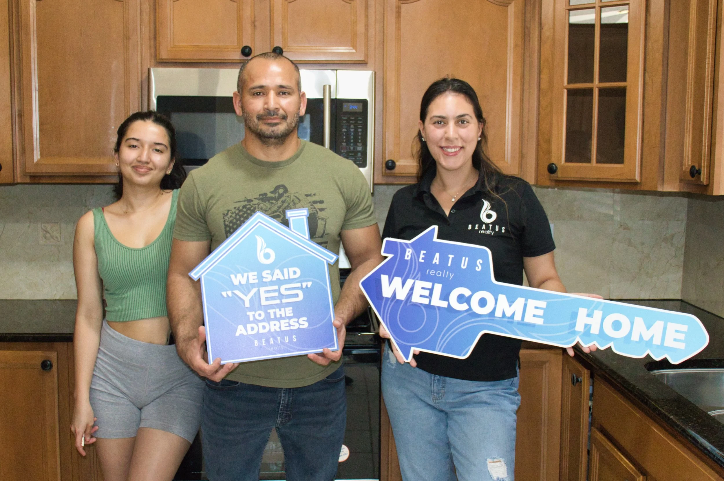 Three people in a kitchen holding signs that say, "We Said 'Yes' to the Address" and "Welcome Home" from Beatus Realty.
