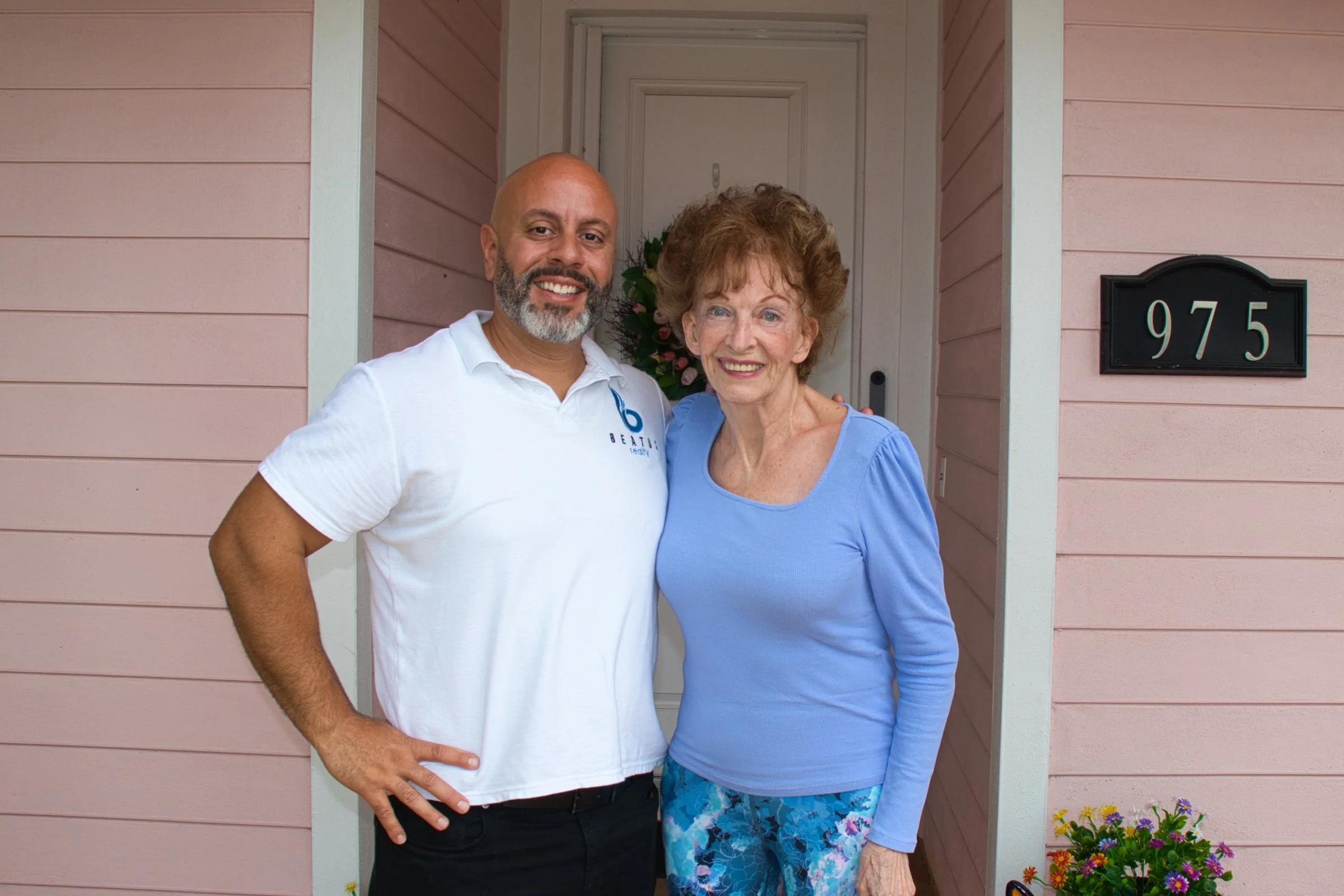 Two people smiling in front of a pink house with the number 975 visible.