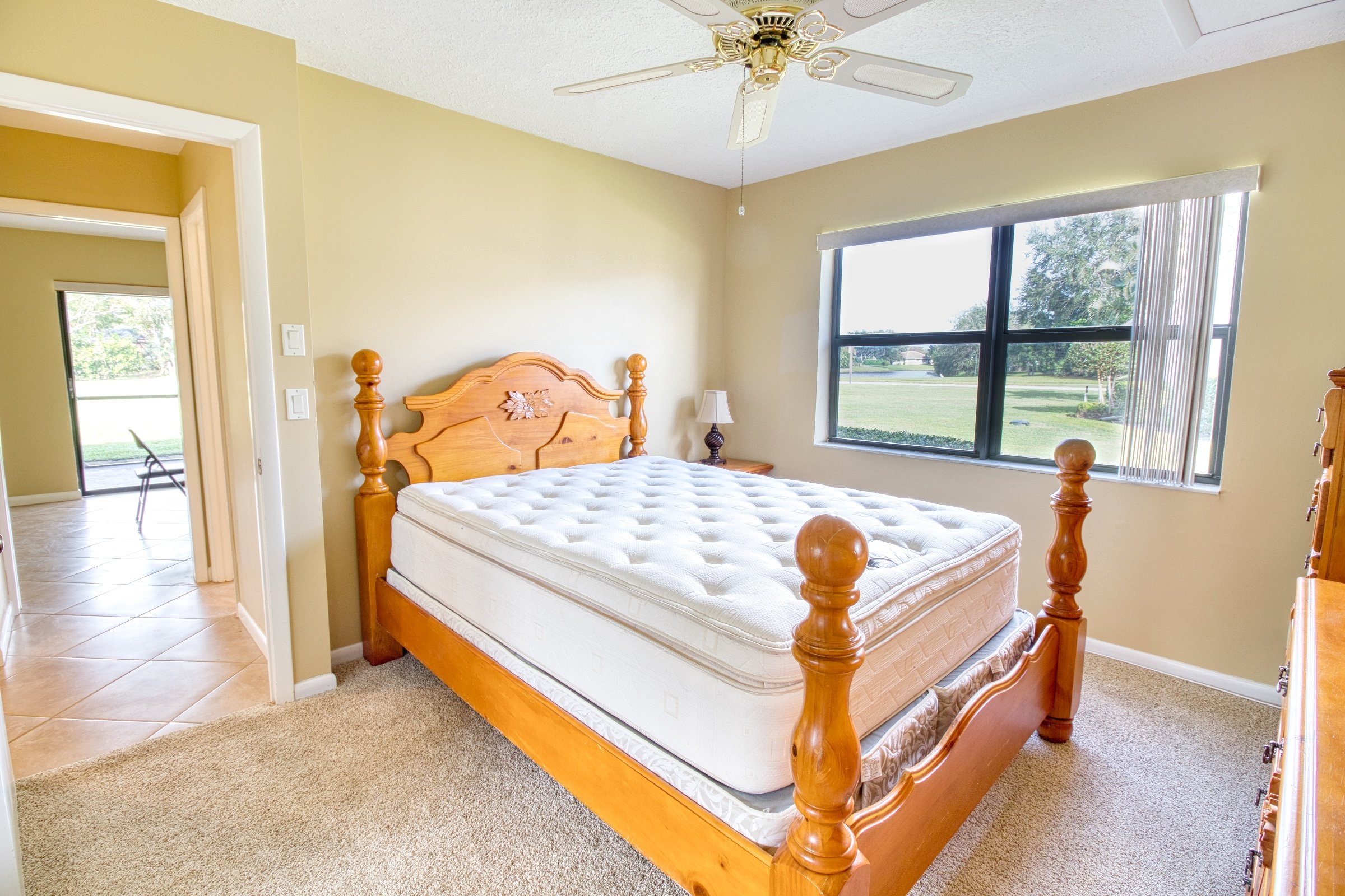 Bedroom with wooden bed frame, white mattress, window view, ceiling fan, bedside lamp, and beige walls.