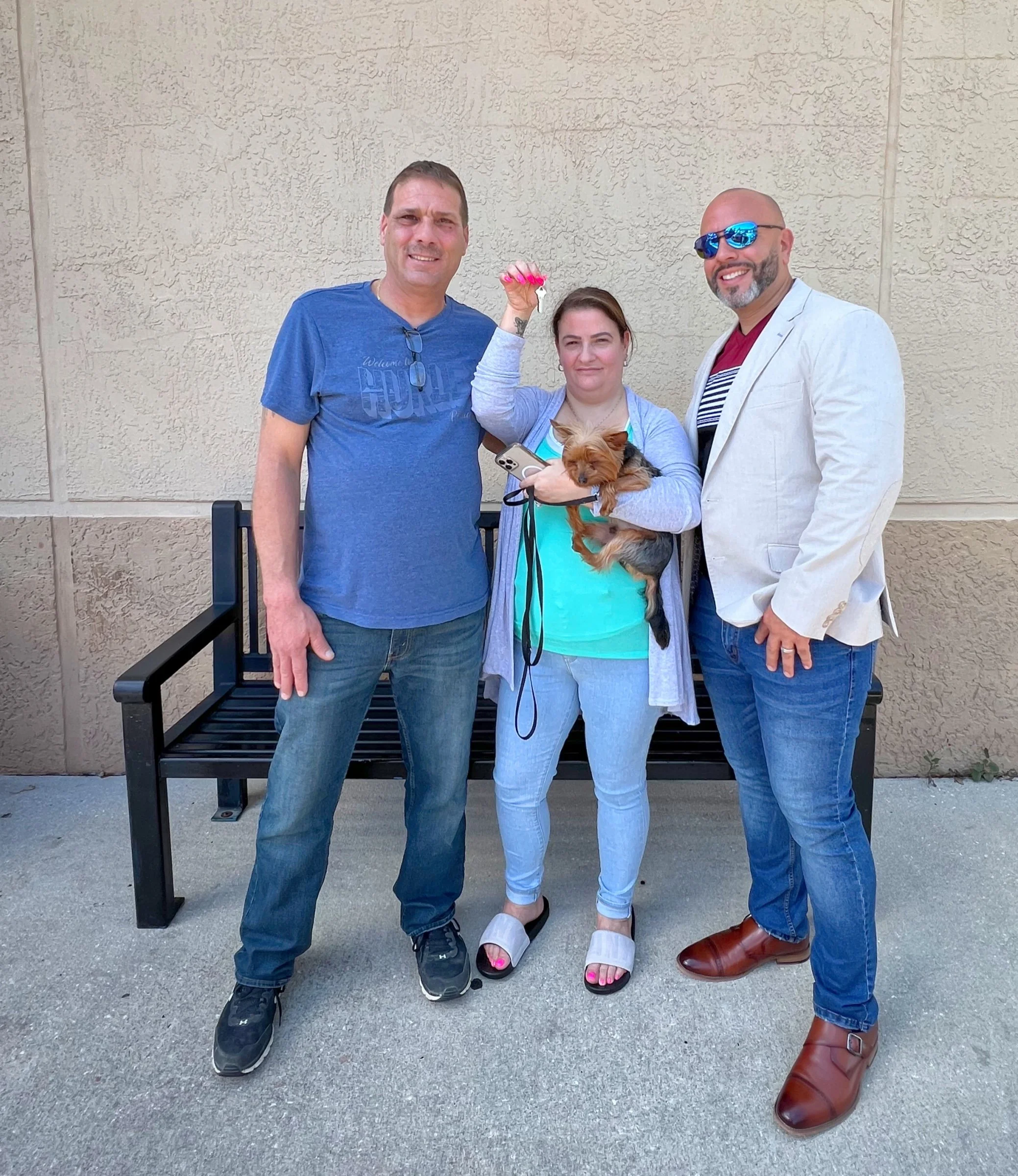 Three people standing outside, two men and a woman holding a small dog, smiling, with a bench in the background.
