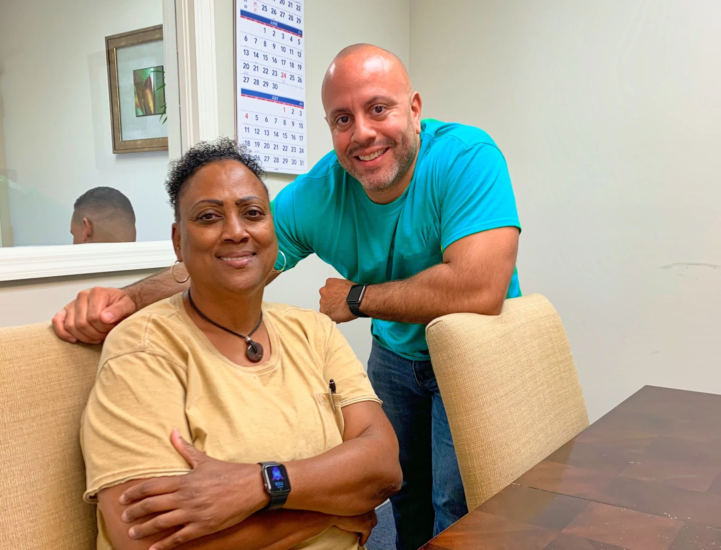 Two people posing in an office setting with a calendar and mirror on the wall.