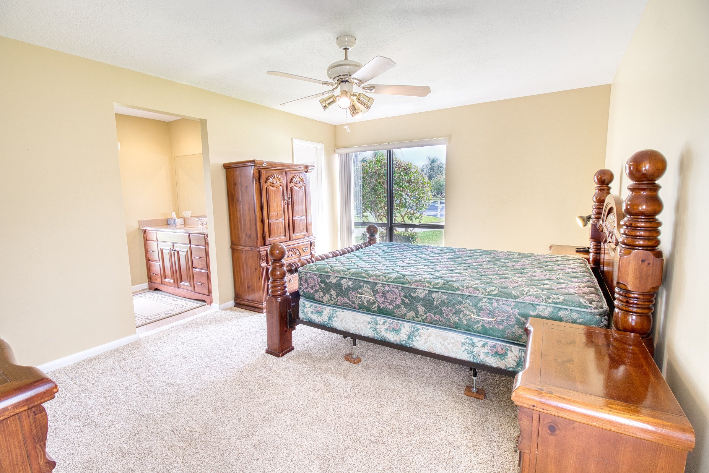 Bedroom with wooden furniture, including a bed with floral mattress, an armoire, nightstands, and a ceiling fan. Room has beige walls, carpeted floor, and a large window with view of a tree.