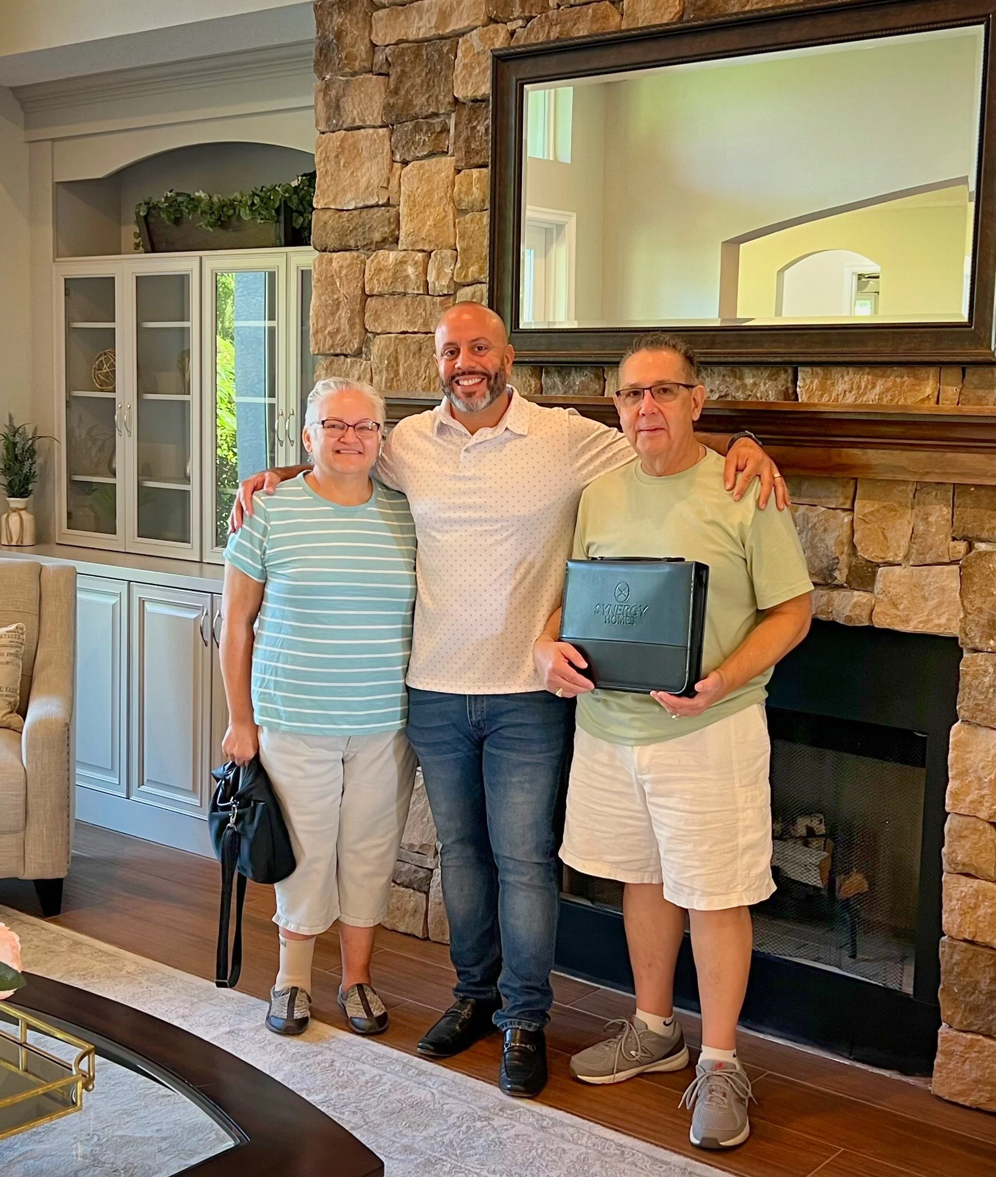 Three people standing indoors in front of a stone fireplace, smiling, one holding a folder. The room is well-lit with a large mirror and cabinets in the background.