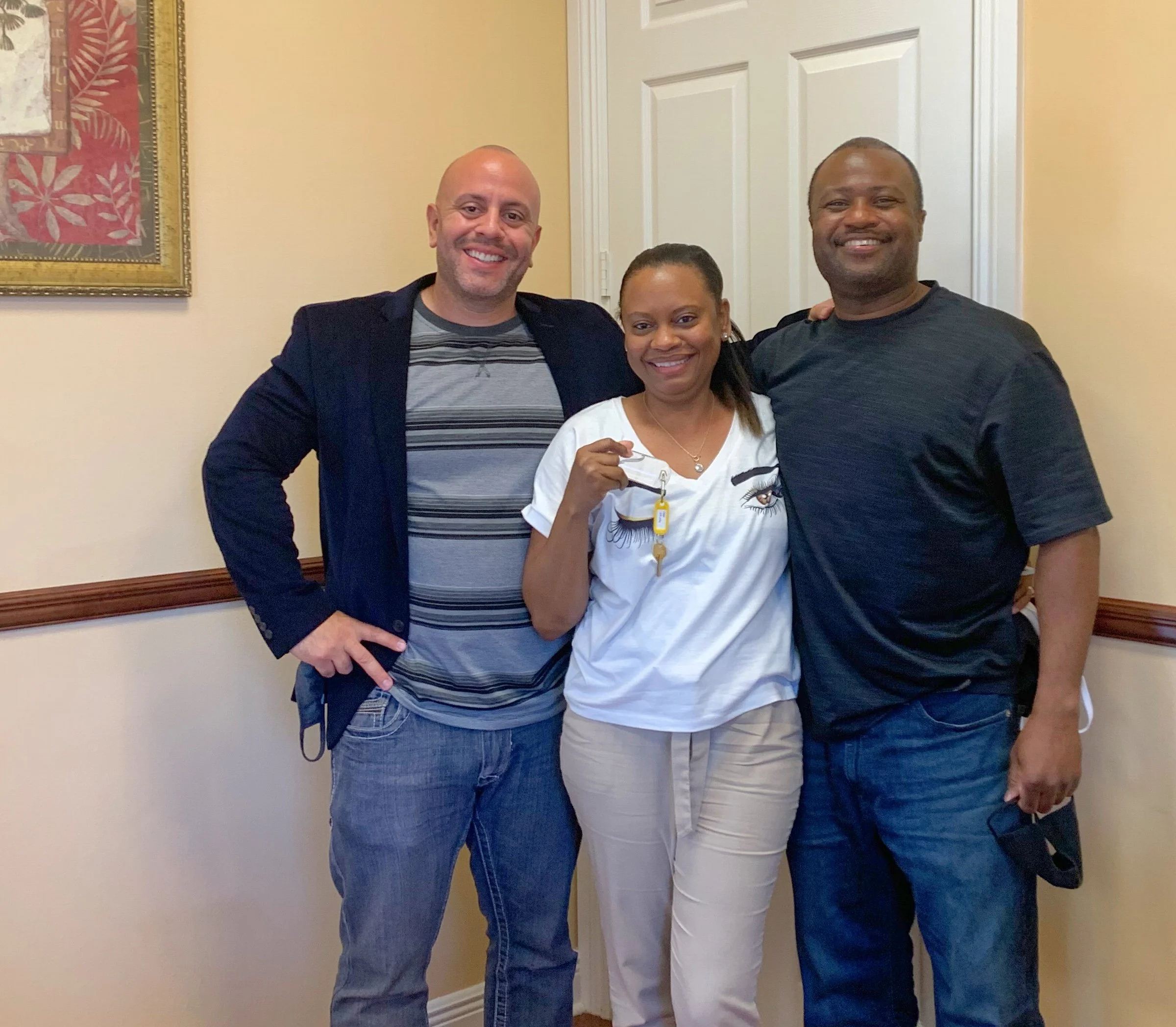 Three people smiling indoors, one woman holding a set of keys, standing between two men.