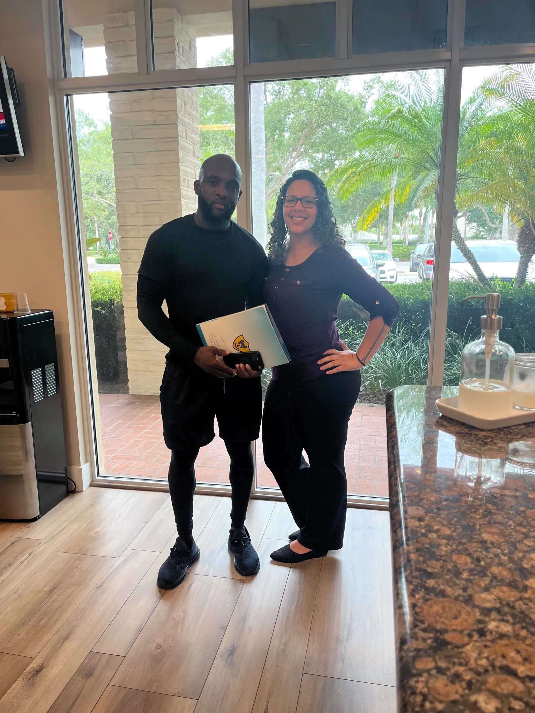 Two people standing indoors near a large window, holding documents, with a view of palm trees and parked cars outside.