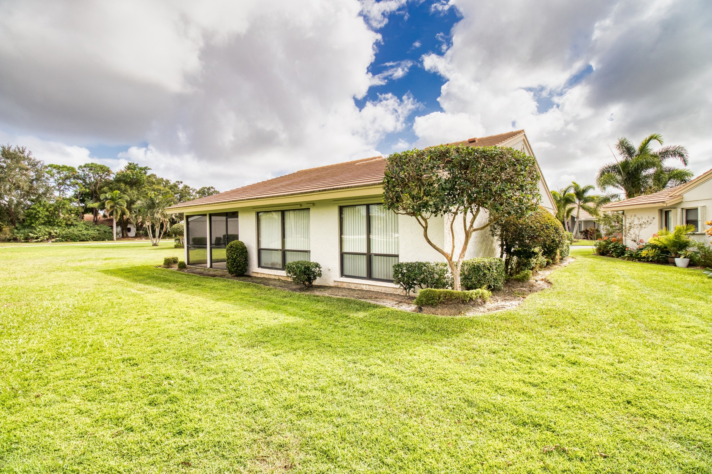 Single-story house with large windows, surrounded by a well-maintained lawn and shrubs, under a cloudy blue sky.
