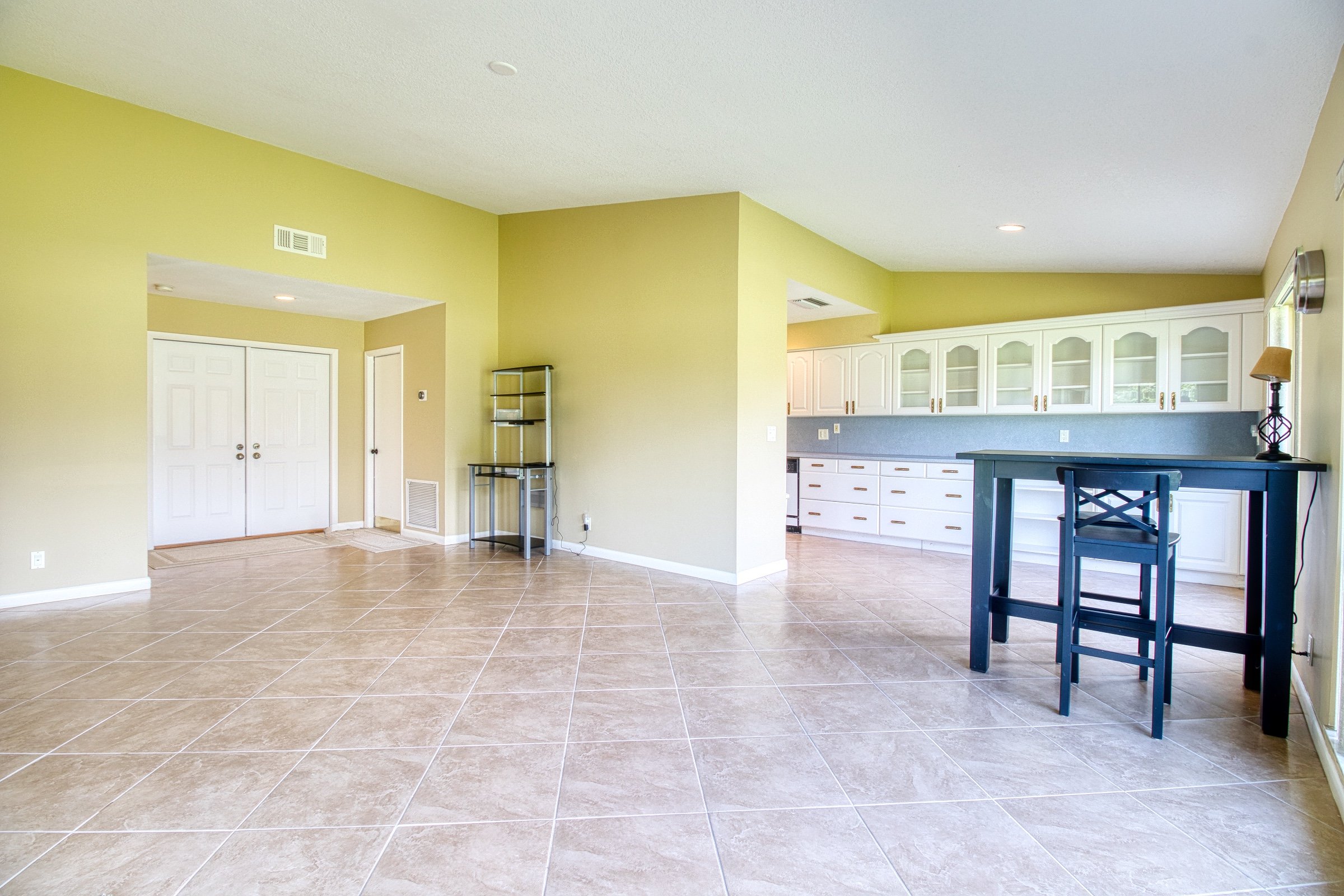Spacious room with tiled flooring and yellow walls, featuring white cabinets, a black table with a chair, and a glass shelf. The room has overhead lighting and a lamp on the table.