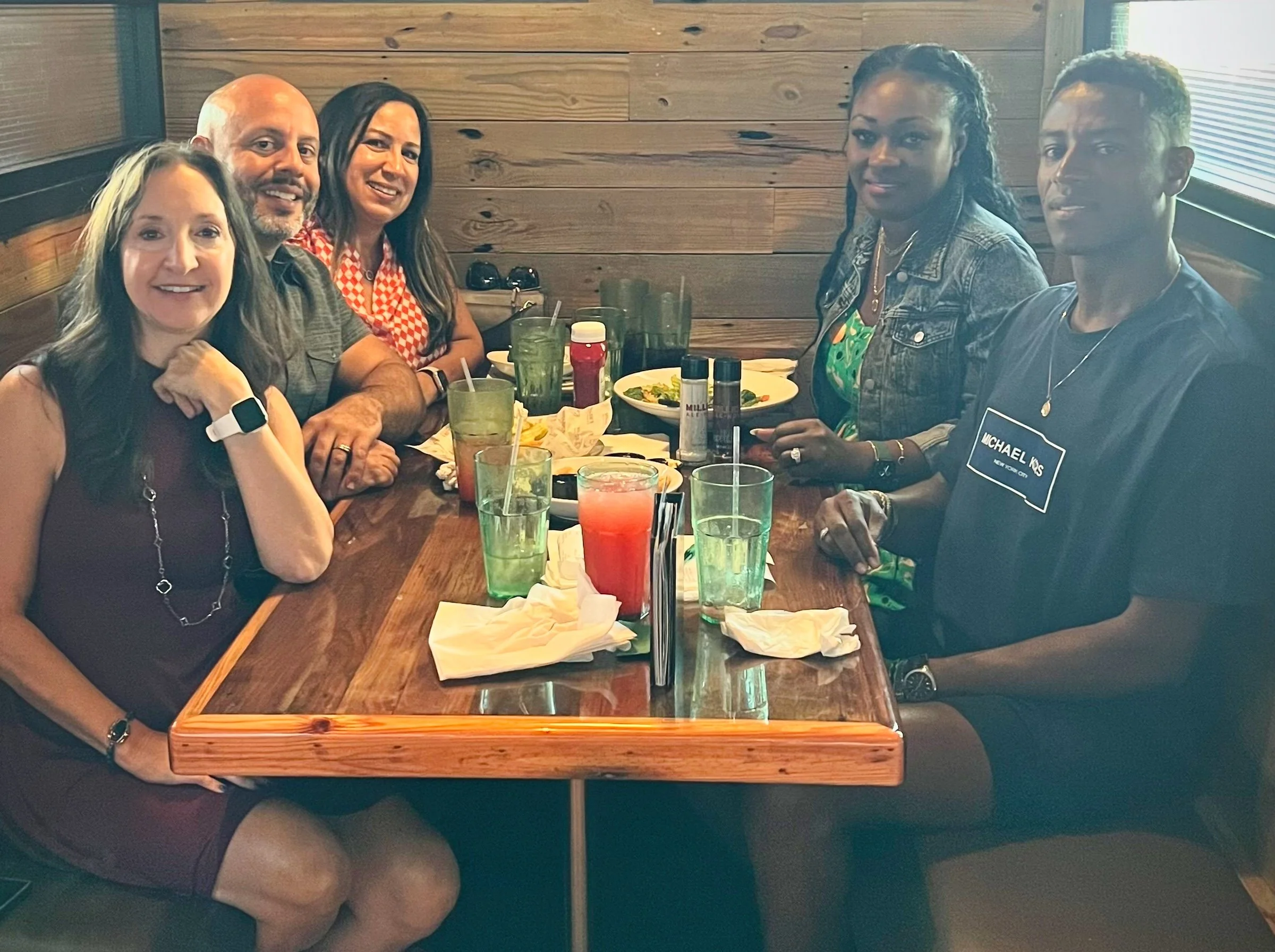 Group of five people dining at a restaurant table with drinks and food; three women and two men sitting closely, smiling, casual attire.
