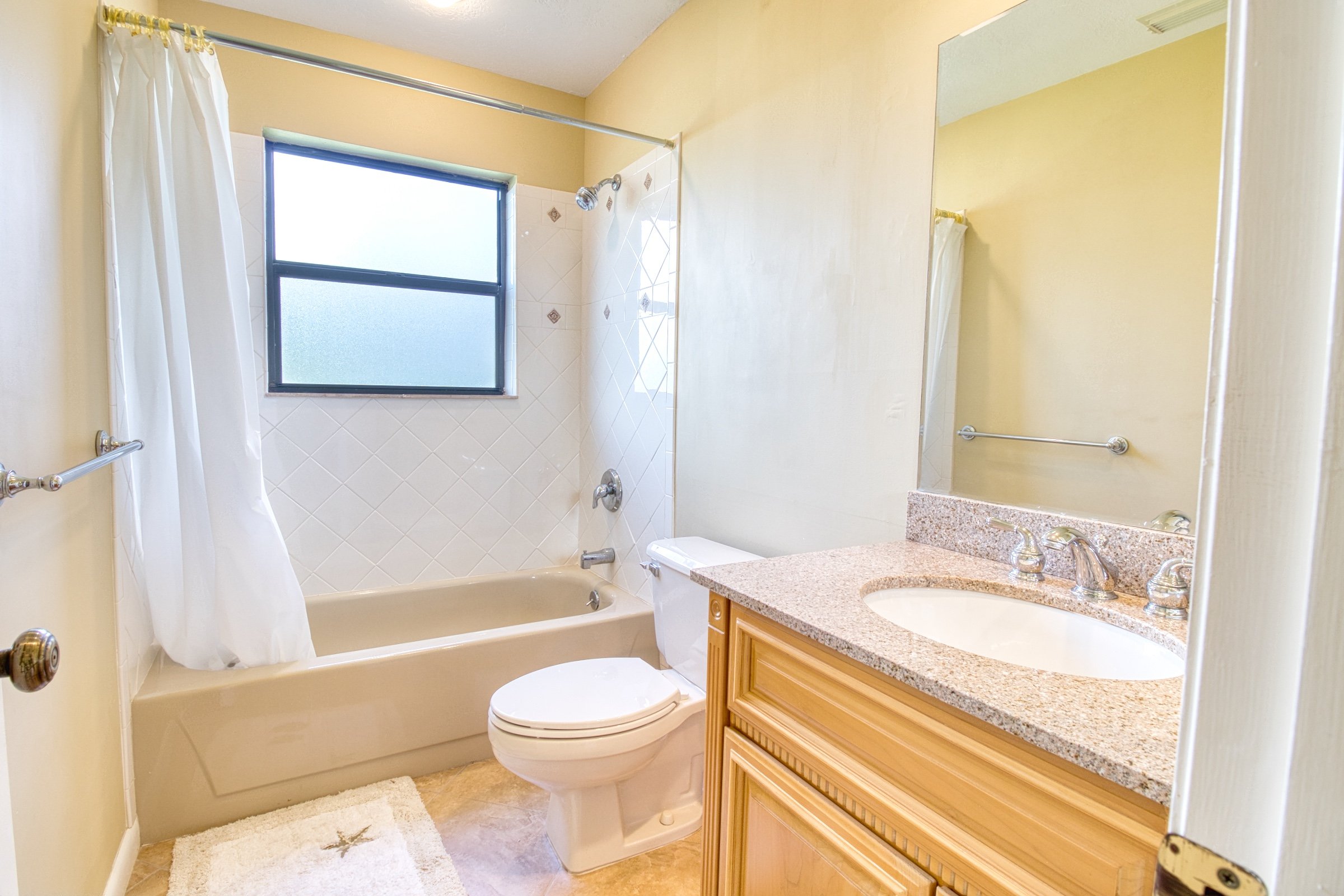 Bathroom with beige bathtub, showerhead, curtain, toilet, granite countertop, wooden vanity, mirror, and window.