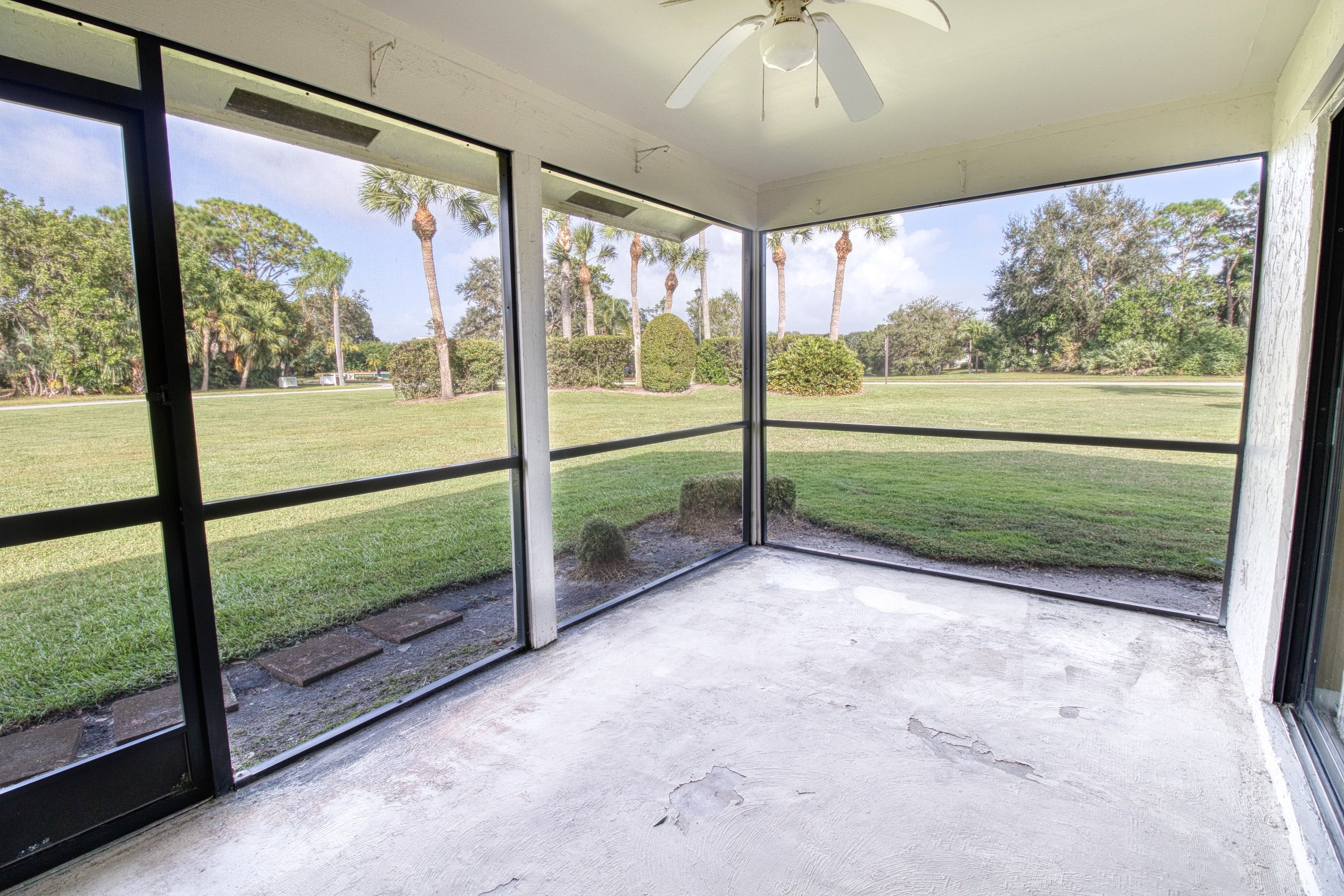 Empty screened porch with ceiling fan, overlooking a green lawn and palm trees.