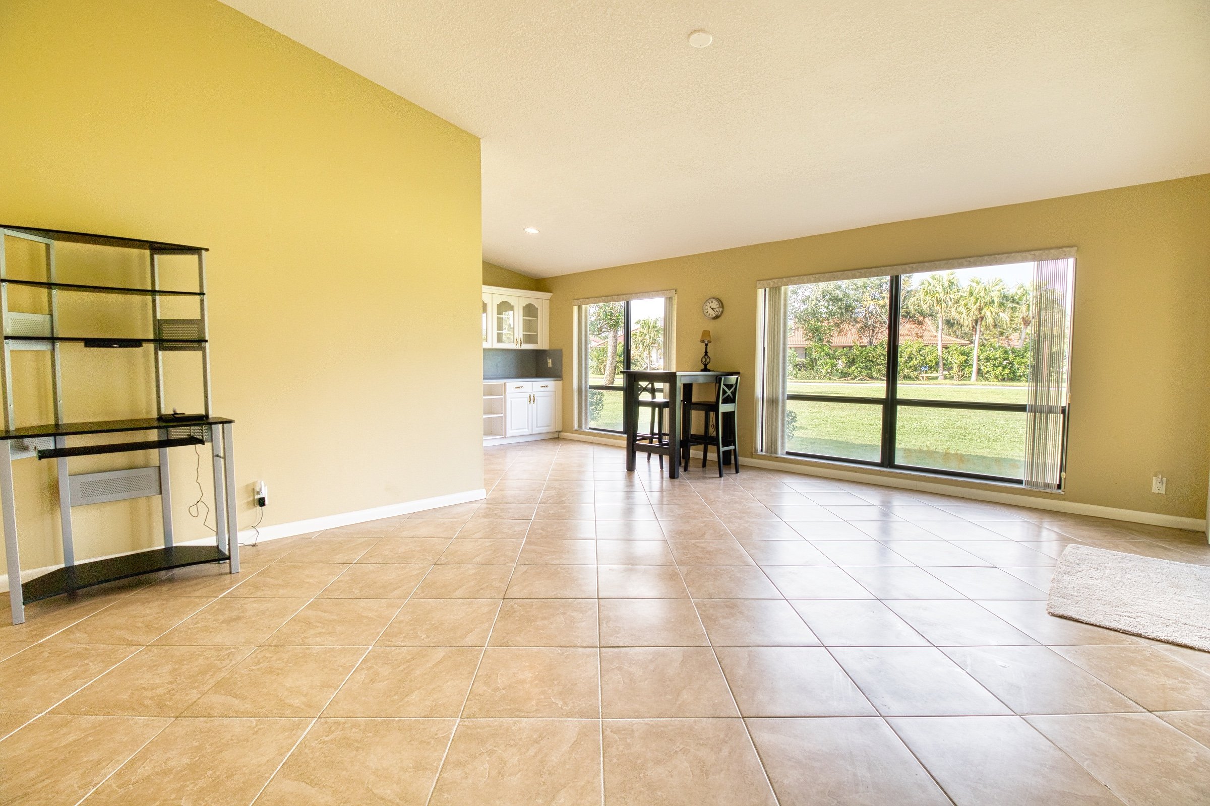 Empty living room with tiled floor, light yellow walls, large windows, black shelving unit, and dining area with two chairs overlooking a grassy exterior.