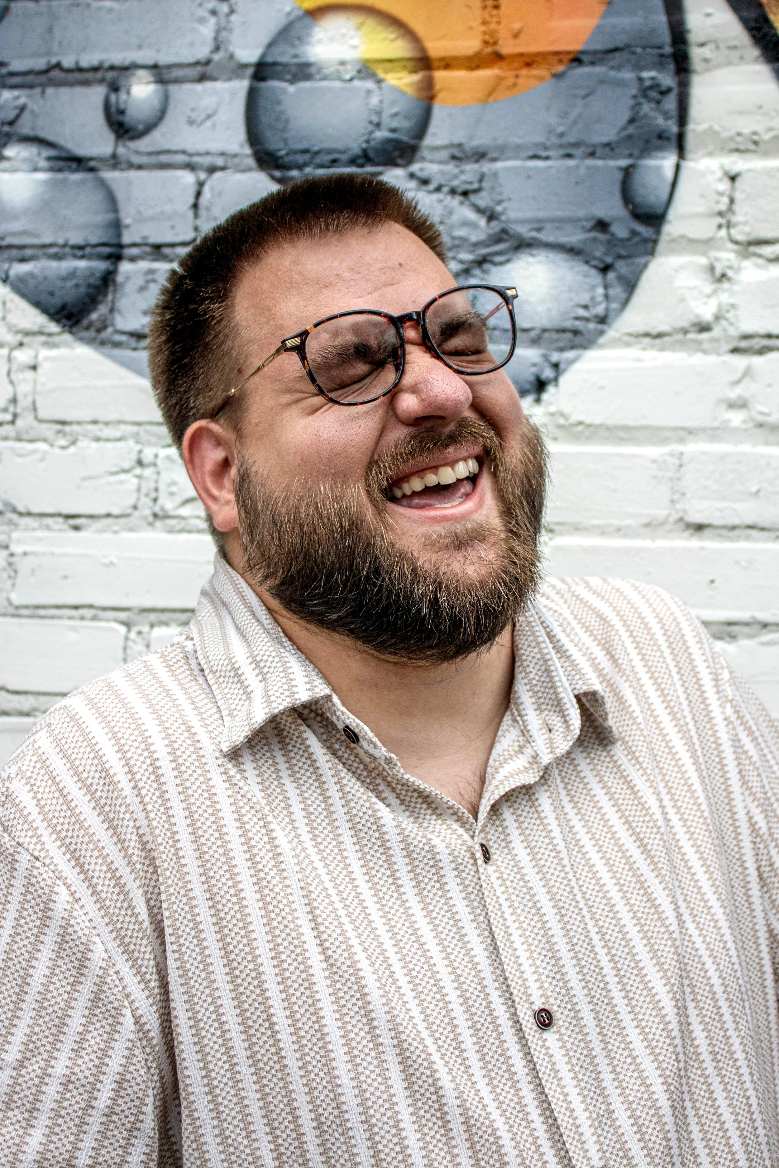 A man with glasses and a beard laughing in front of a white brick wall with a heart-shaped mural.