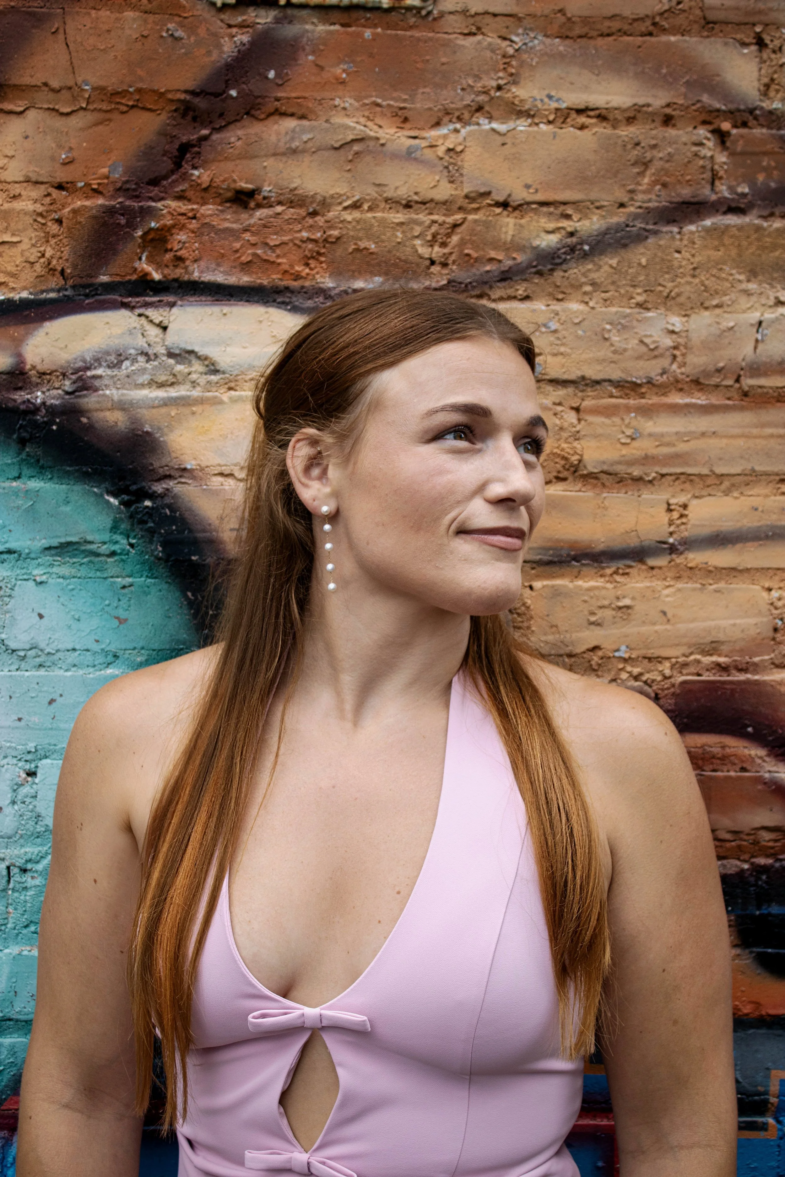 A woman with long red hair wearing a pink sleeveless top with cutout and bow details, standing against a brick wall with graffiti.