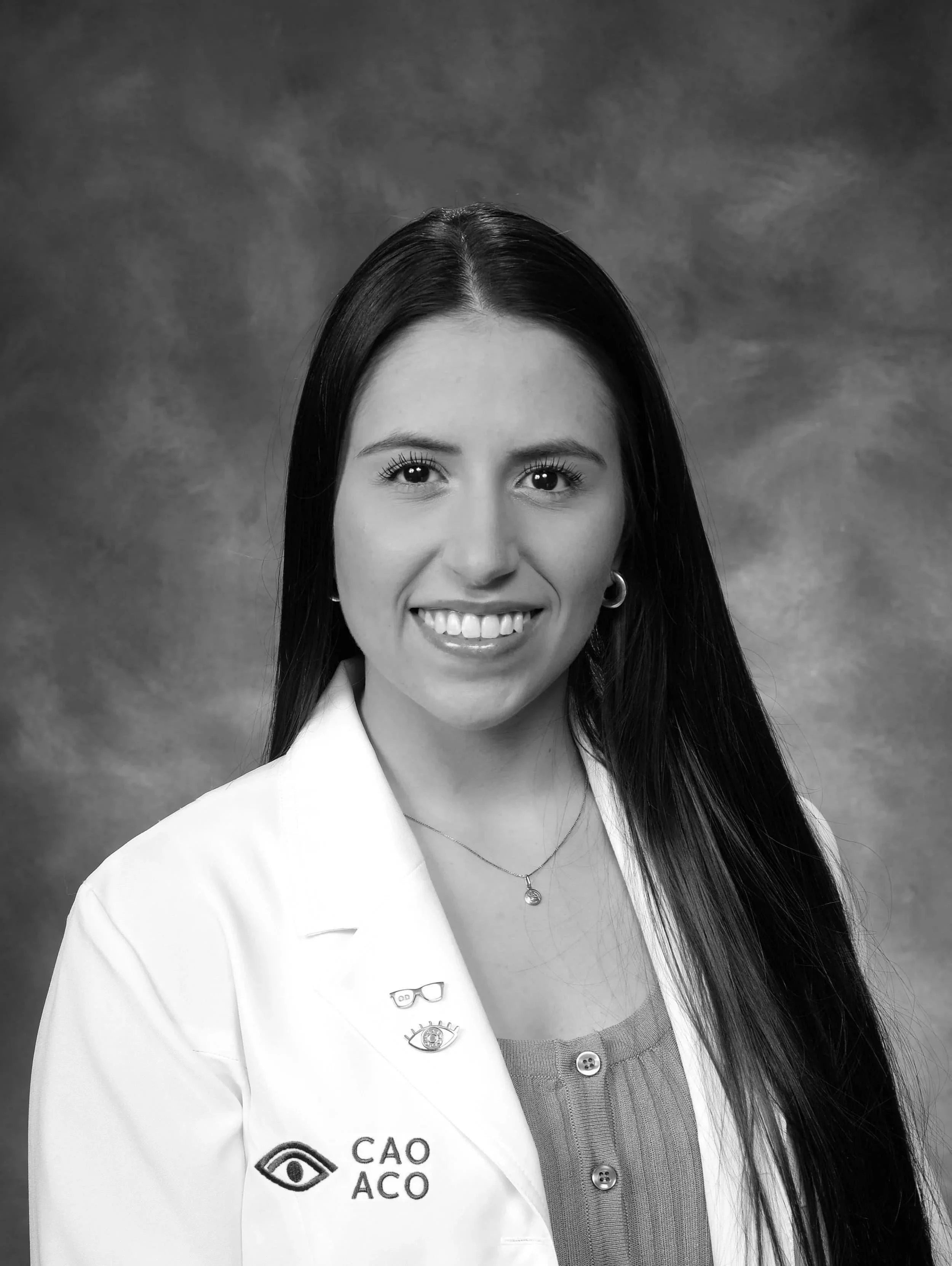 A woman with long dark hair smiles, wearing a white lab coat and necklace, in front of a studio backdrop.