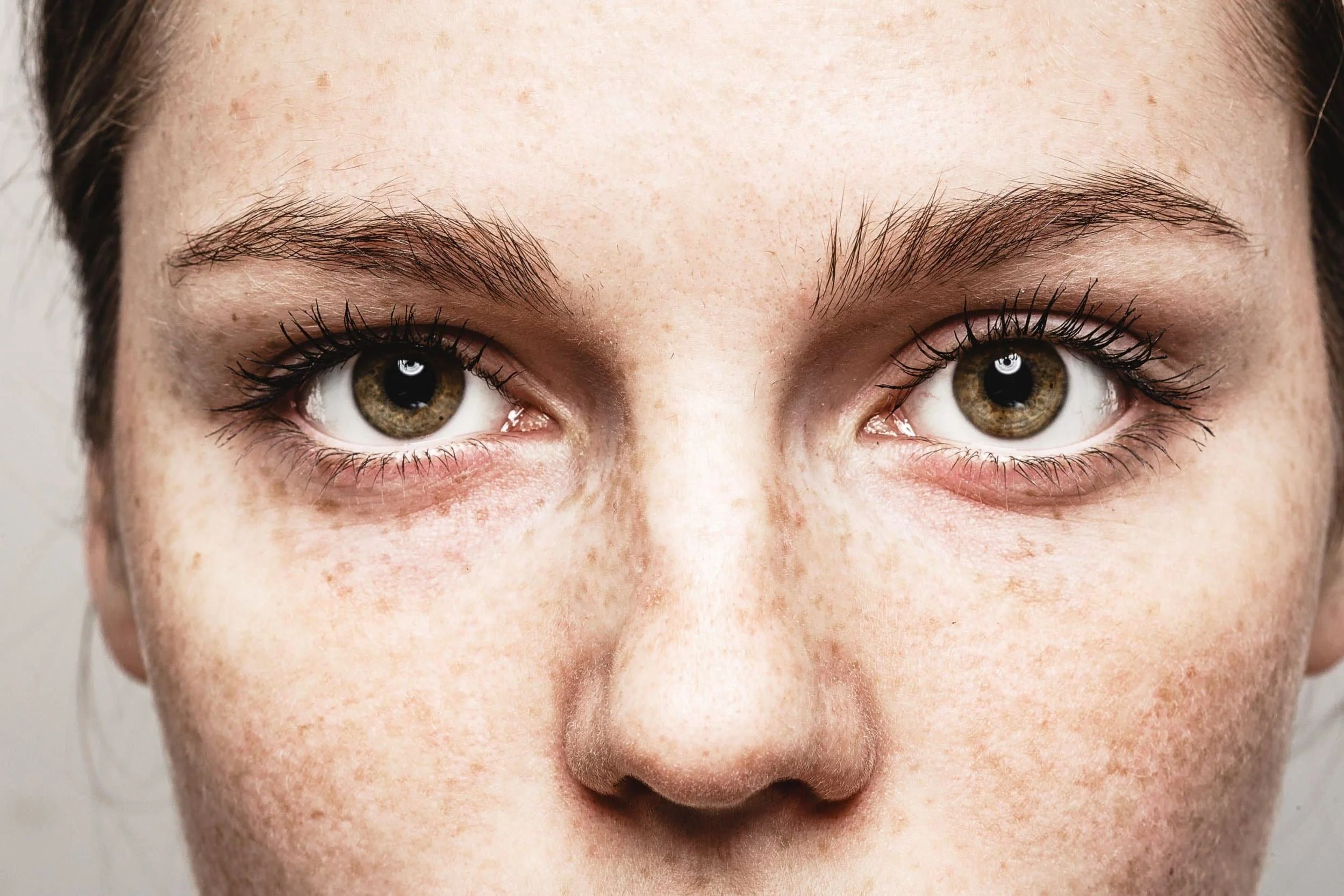 Close-up of a persons face, focusing on greenish-brown eyes, eyebrows, and freckles on fair skin.