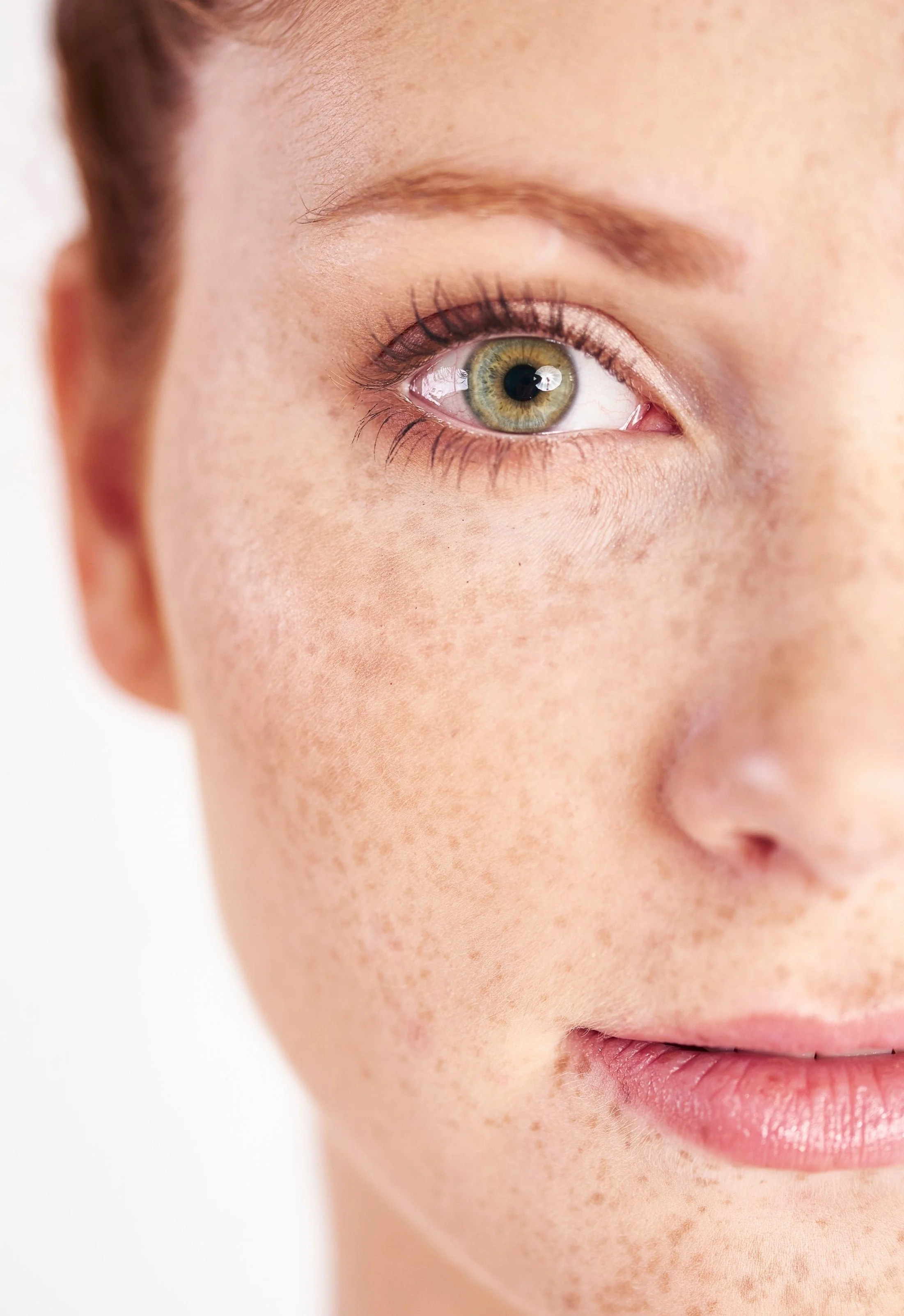 Gros plan d'un visage féminin, mettant en évidence un œil vert, des tâches de rousseur sur la joue et des lèvres roses.