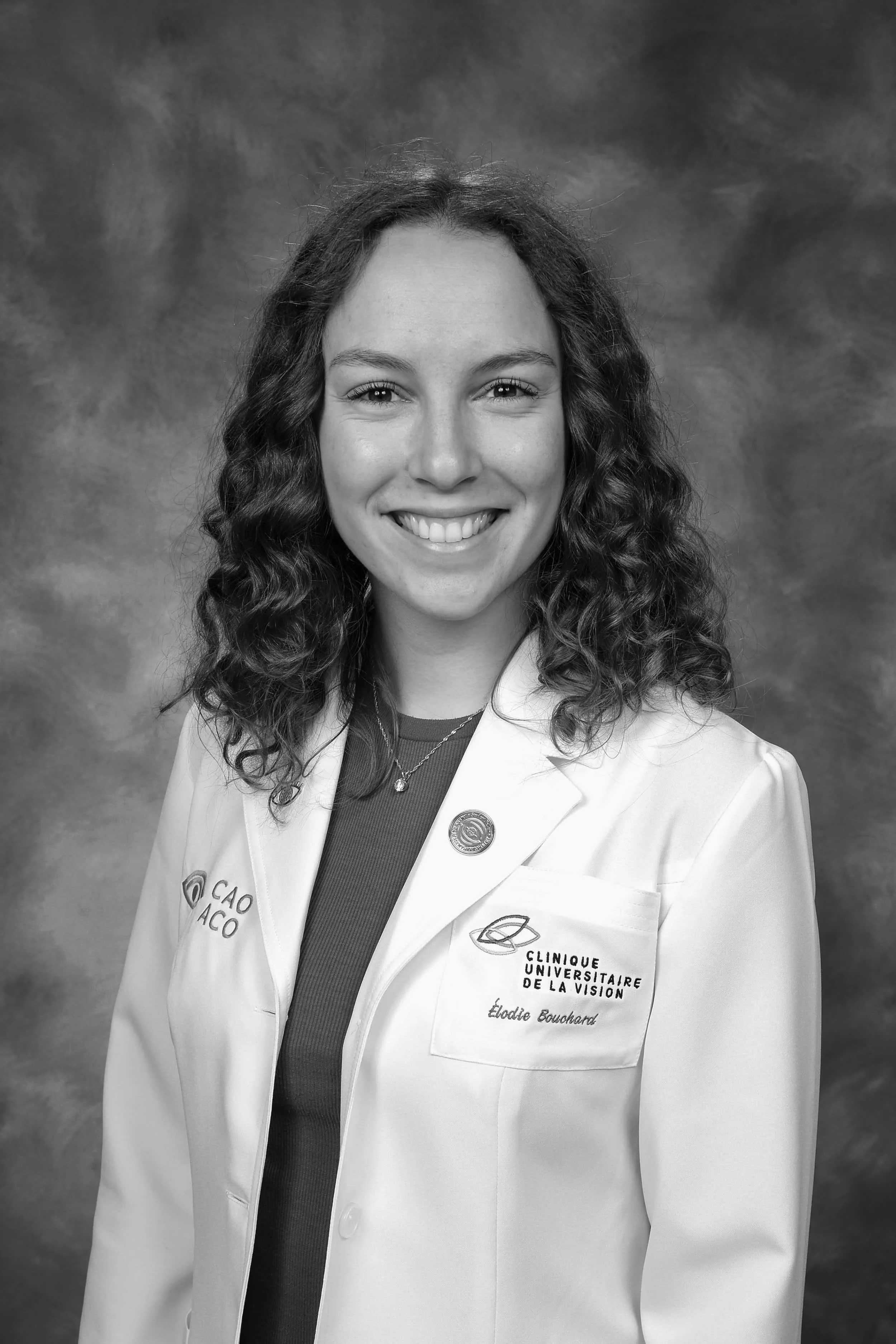 Smiling woman with curly hair in a white lab coat, standing in front of a studio backdrop.