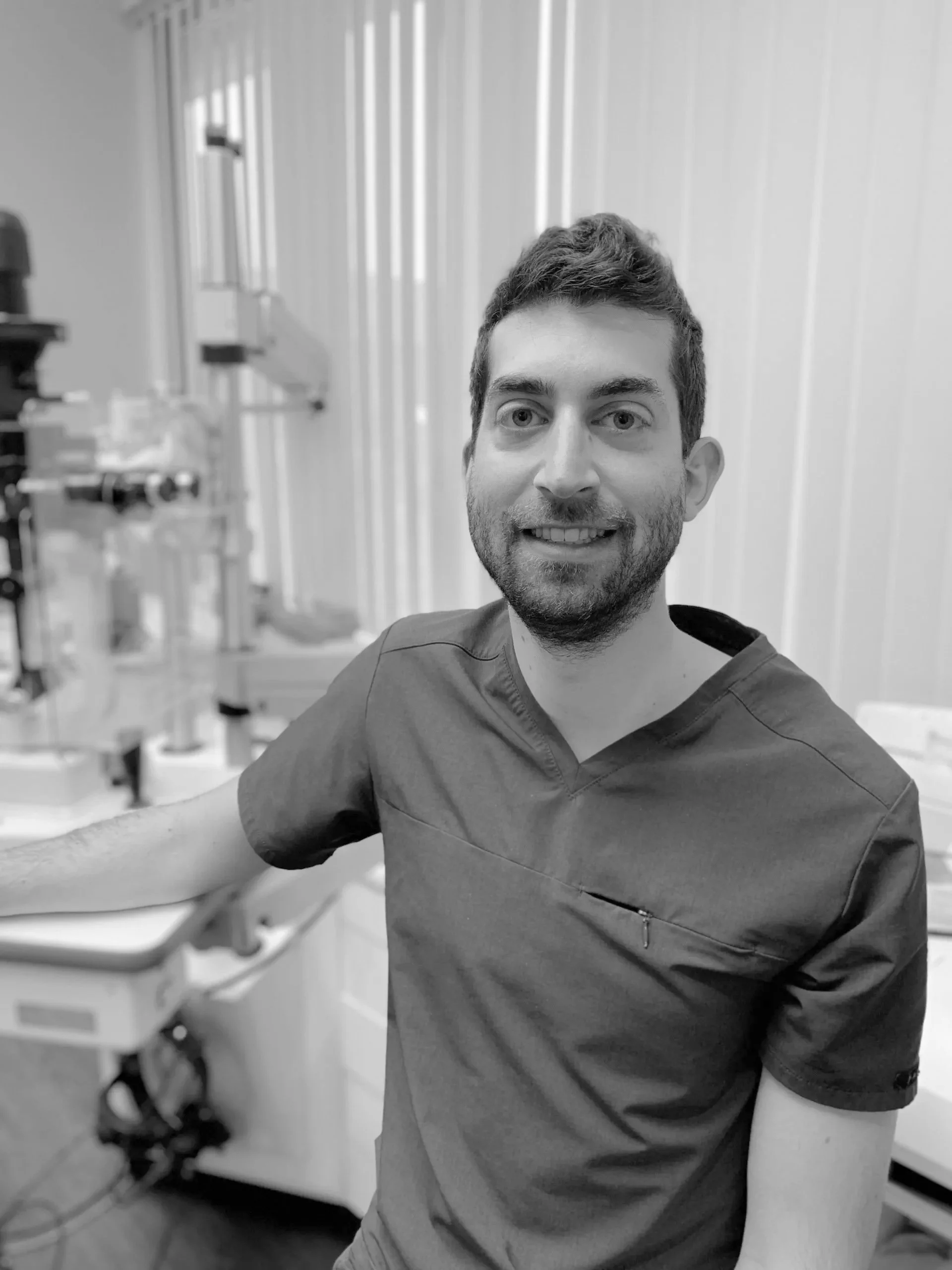A man in medical scrubs smiles while seated in a clinic or lab with medical equipment in the background.
