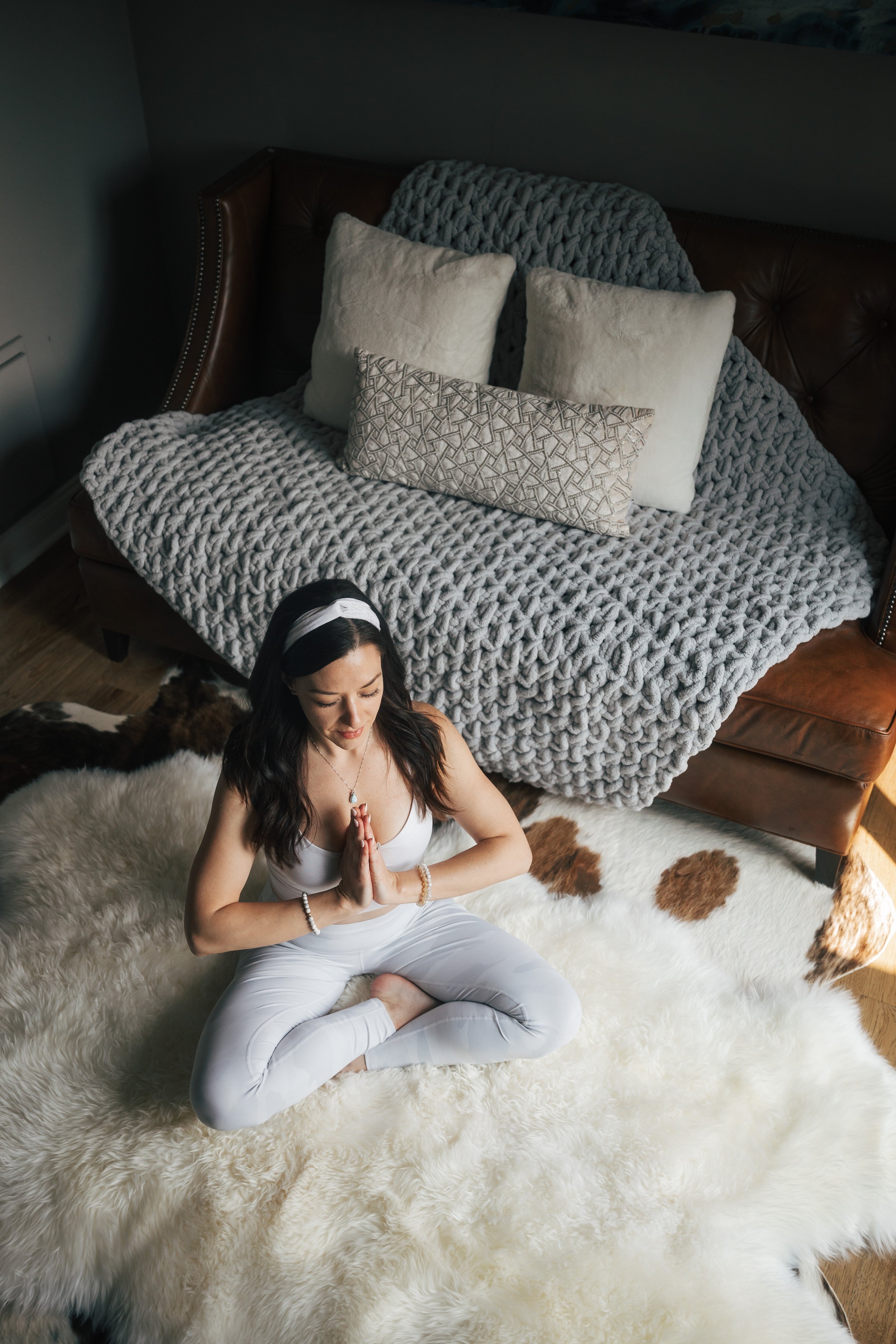 A woman practicing yoga or meditation sitting cross-legged on a fluffy white rug in a living room, with hands in prayer position, head bowed, and eyes closed.