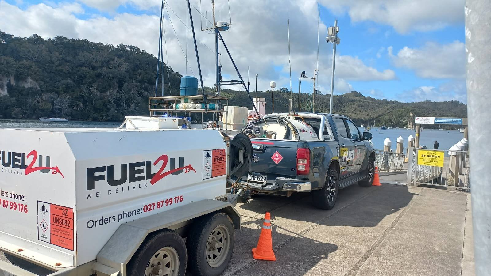 A boat being refueled with a fuel truck at a harbor with water and boats in the background, surrounded by a secure gate with a yellow sign and orange cones.