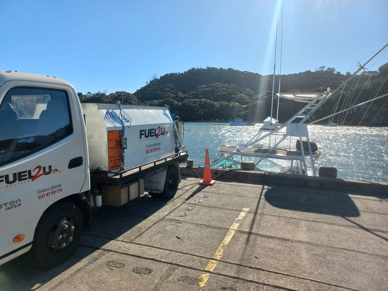 A boat is docked next to a marina in a scenic harbor, with a fuel delivery truck parked nearby on a concrete pier, and a hillside covered in trees in the background.