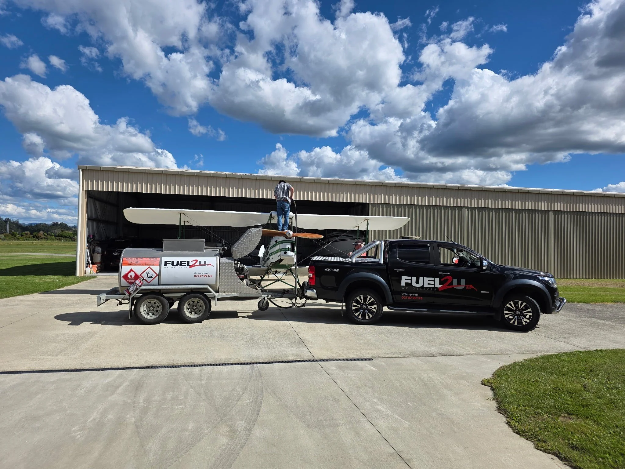 A man standing on a small aircraft on a trailer attached to a black pickup truck in front of a large metal building, with a partly cloudy sky overhead.