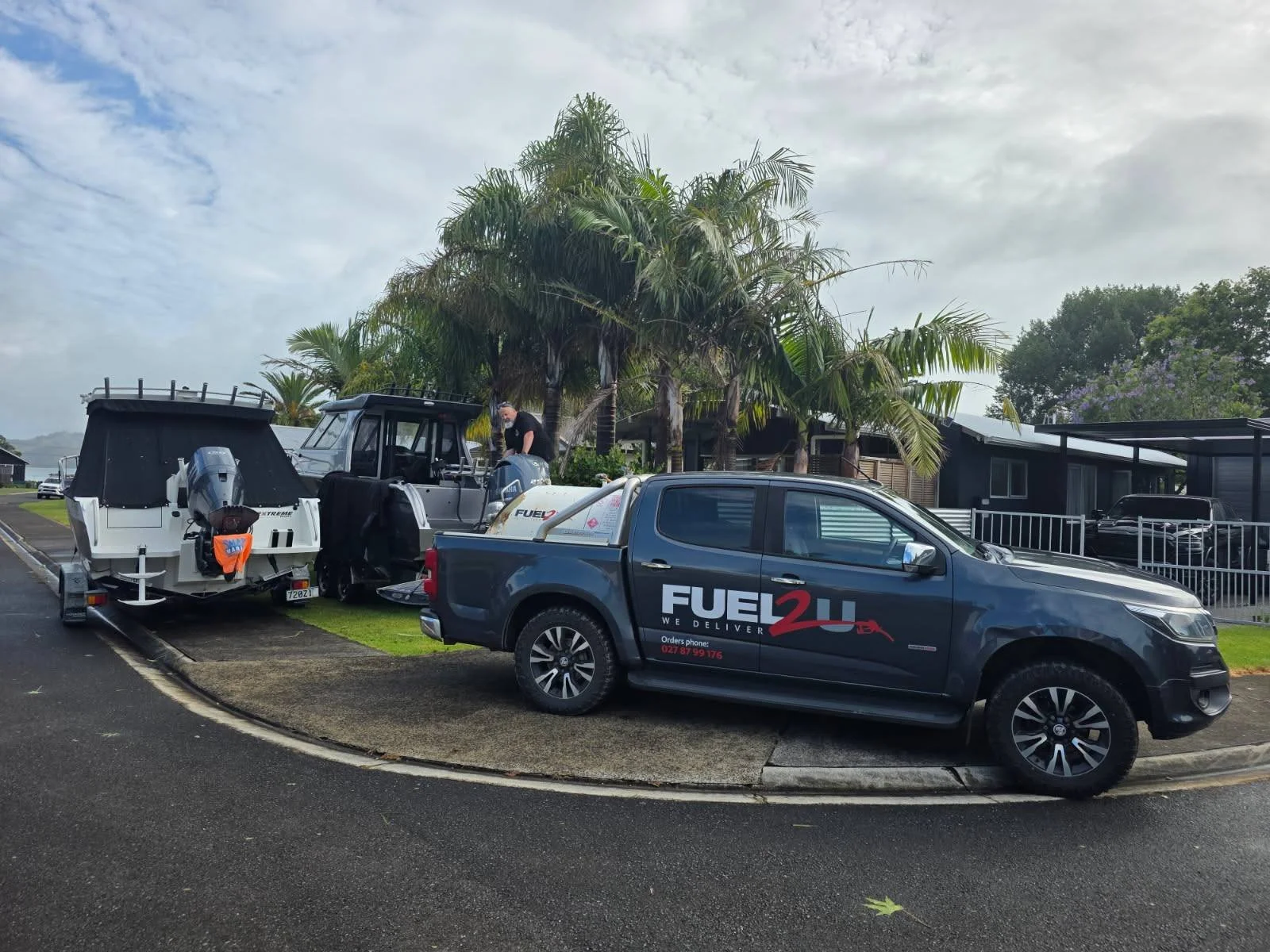 A black pickup truck with a «Fuel2U» logo parked on the side of a street. Behind it, there are boats, one on a trailer and one on a stand, with a person working on or near the boat. There are palm trees and houses in the background under a cloudy sky