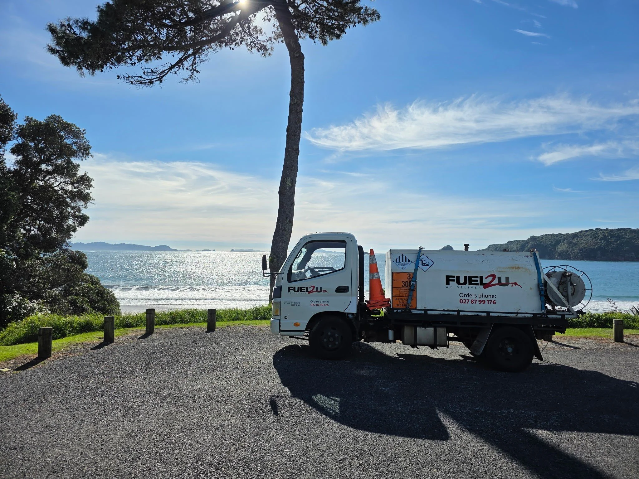 A white service truck with orange safety cones in a parking lot near a scenic coastal area with water and mountains in the distance. There is a tall tree and a clear blue sky with some wispy clouds.