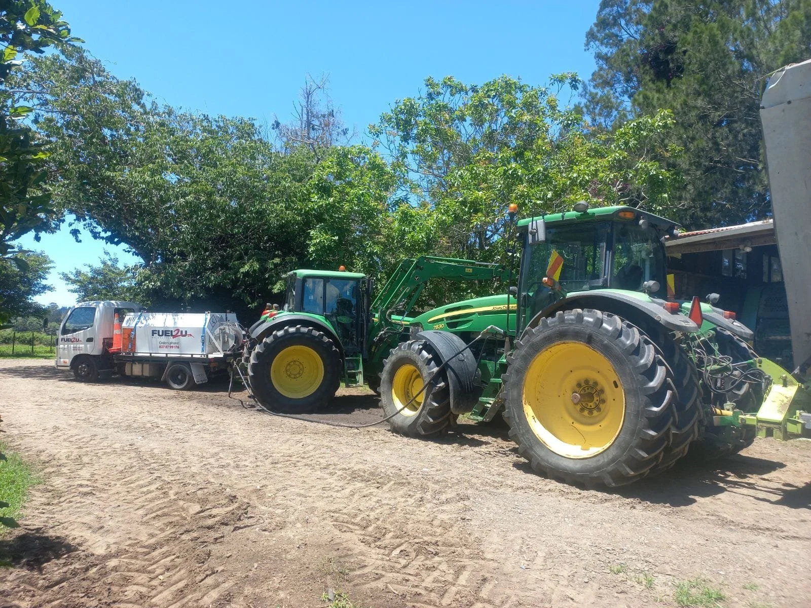 A large green John Deere tractor connected to a fuel truck on a sandy outdoor area, surrounded by trees and blue sky.