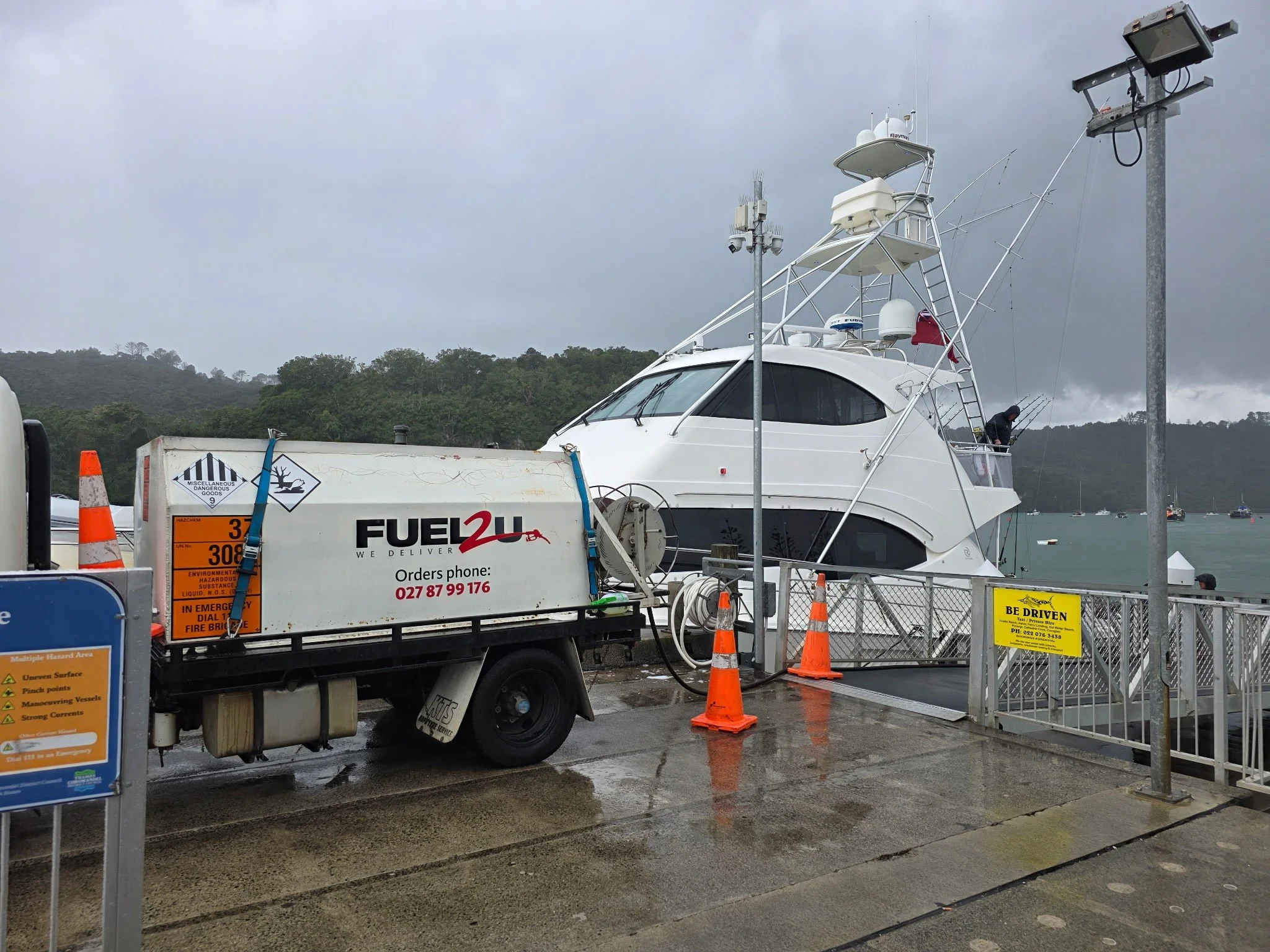 A white yacht docked at a marina under cloudy skies, with various fishing rods on the deck. In the foreground, there is a fuel truck labeled Fuel 2U, surrounded by orange safety cones. The scene appears wet, possibly after rain, with hills and boats 