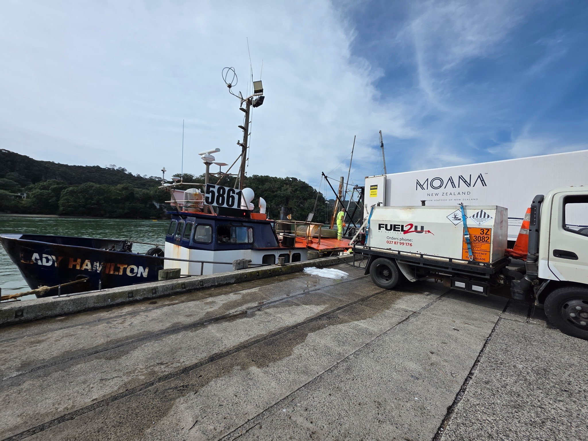 A boat docked at a pier near a shoreline with trees, with a trailer truck nearby carrying equipment for seafood delivery.