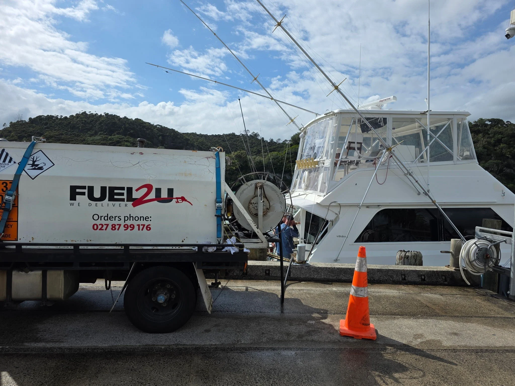 A boat being refueled at a fueling station, with orange cones and a Fuel 2U fuel truck nearby, against a background of hillside with trees and a partly cloudy sky.
