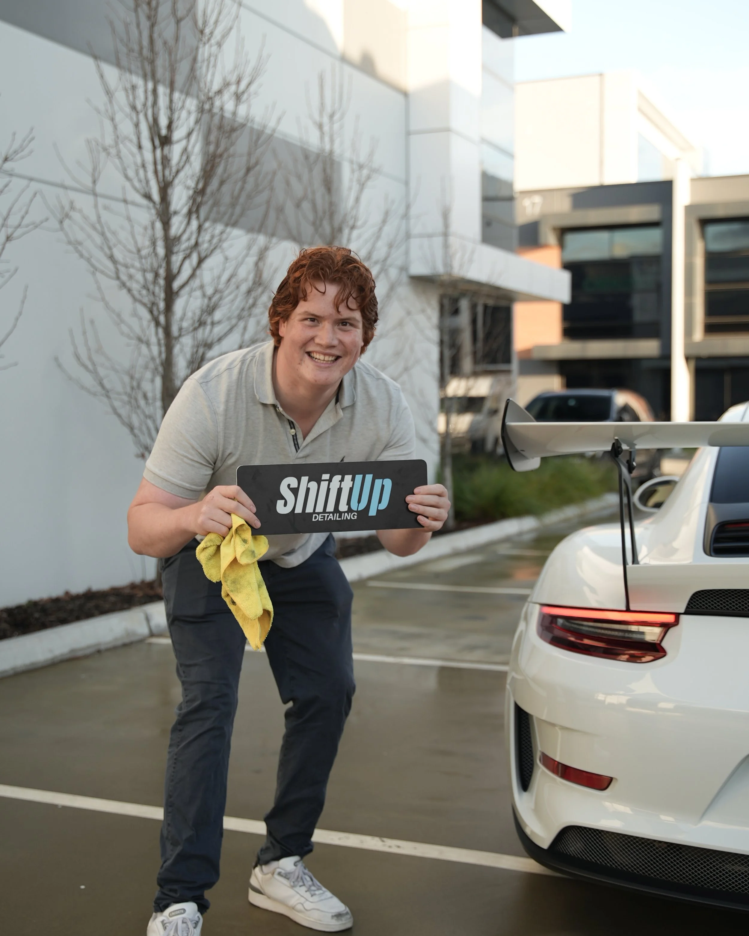 Smiling young man with curly red hair holding a black and white sign that reads 'Shift Up Detailing', standing next to a white sports car with its rear and rear wing visible, in an outdoor parking lot with modern white and black buildings in the background.