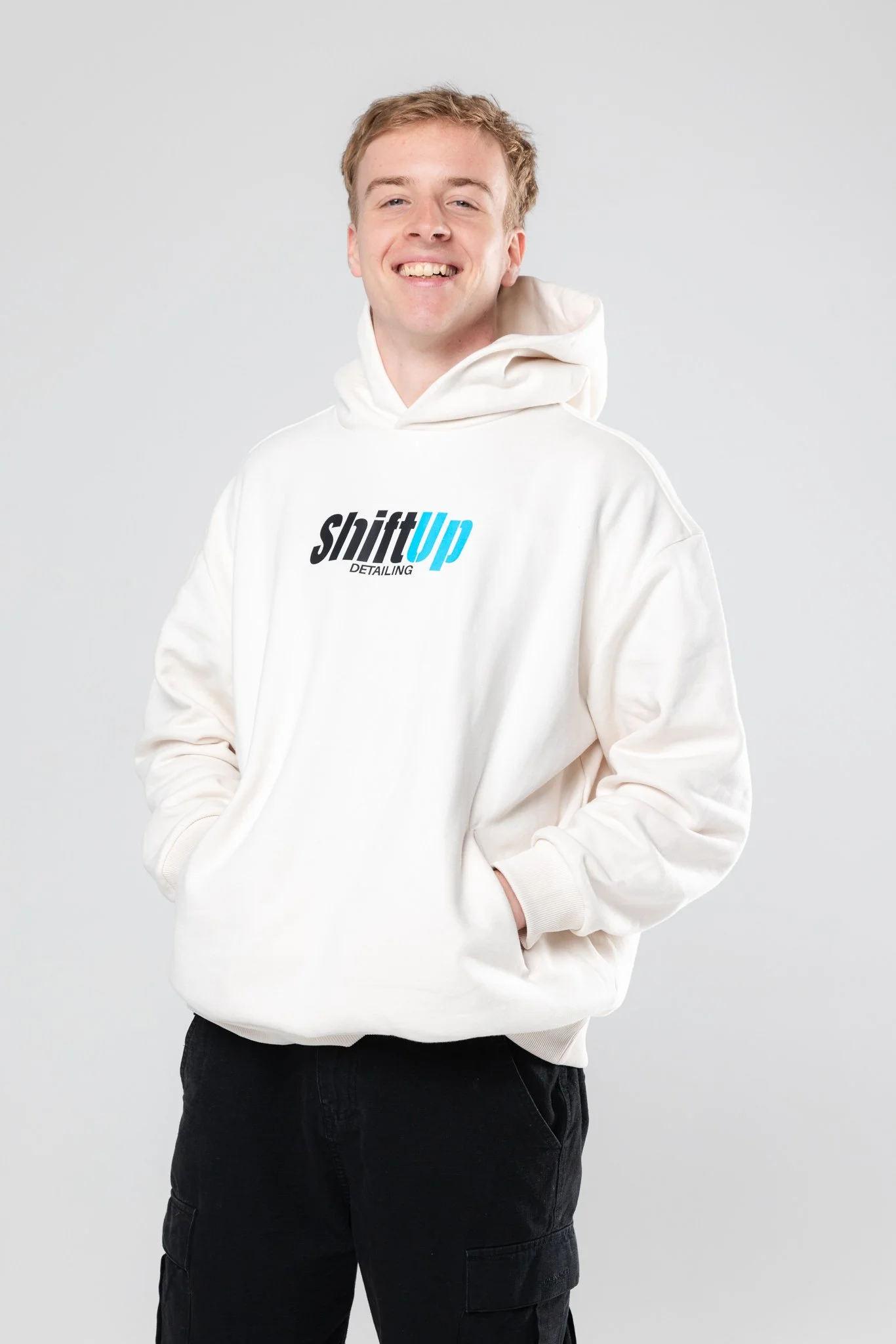 Jacob lewis smiling, wearing a white hoodie with 'shiftUp Detailing' logo, black pants, standing against a plain gray background.