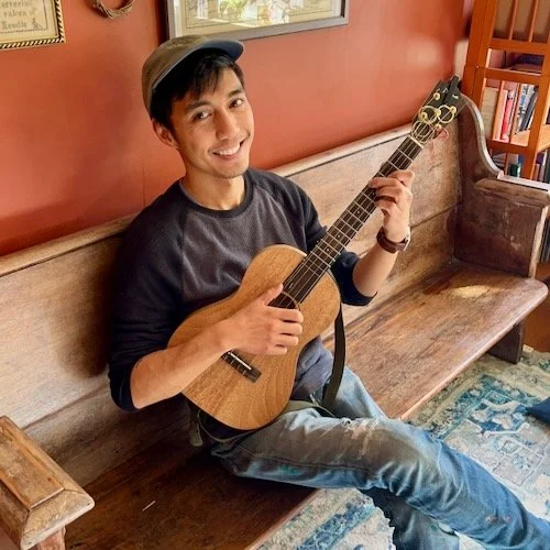 armand aromin sitting on antique pew with ukelele smiling at the camera