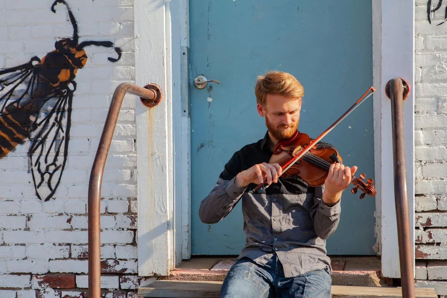 Oliver Scalon in front of a blue door sitting on stoop playing a fiddle. 