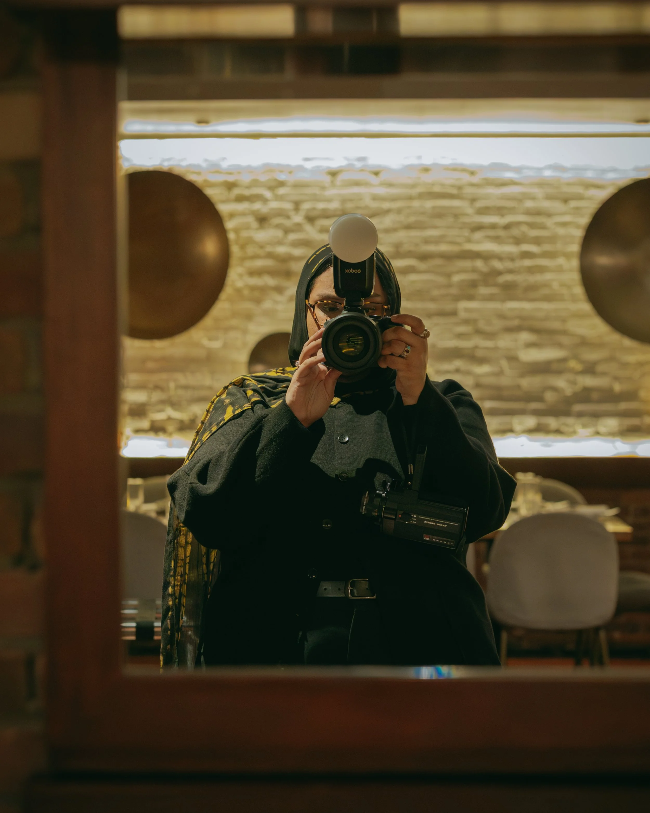 Person taking a photo in a mirror with a camera, in a room with brick walls and round wall decorations.