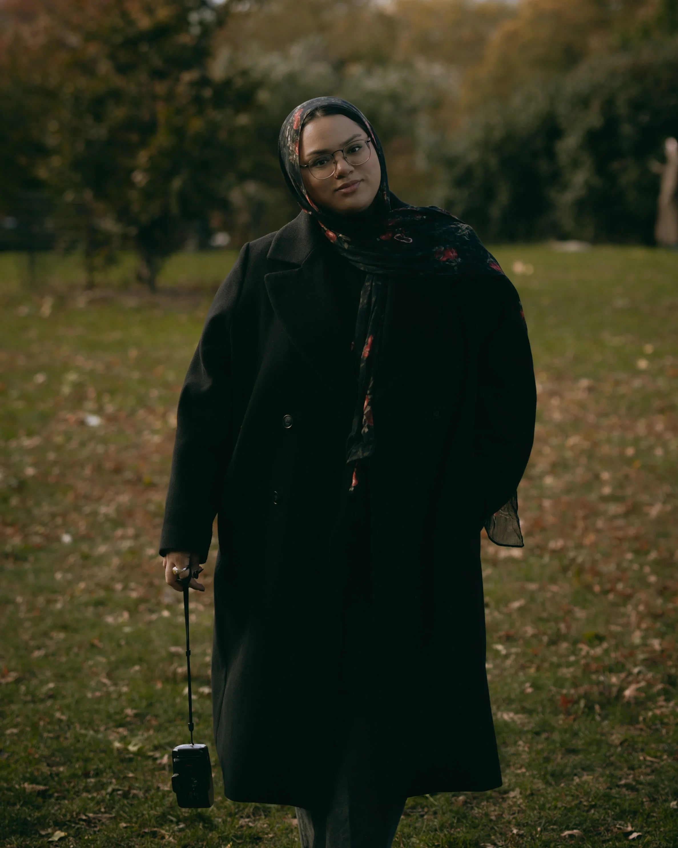 A woman standing outdoors in a park with autumn leaves, wearing glasses, a floral headscarf, and a long black coat, holding a small camera on a strap in her right hand.