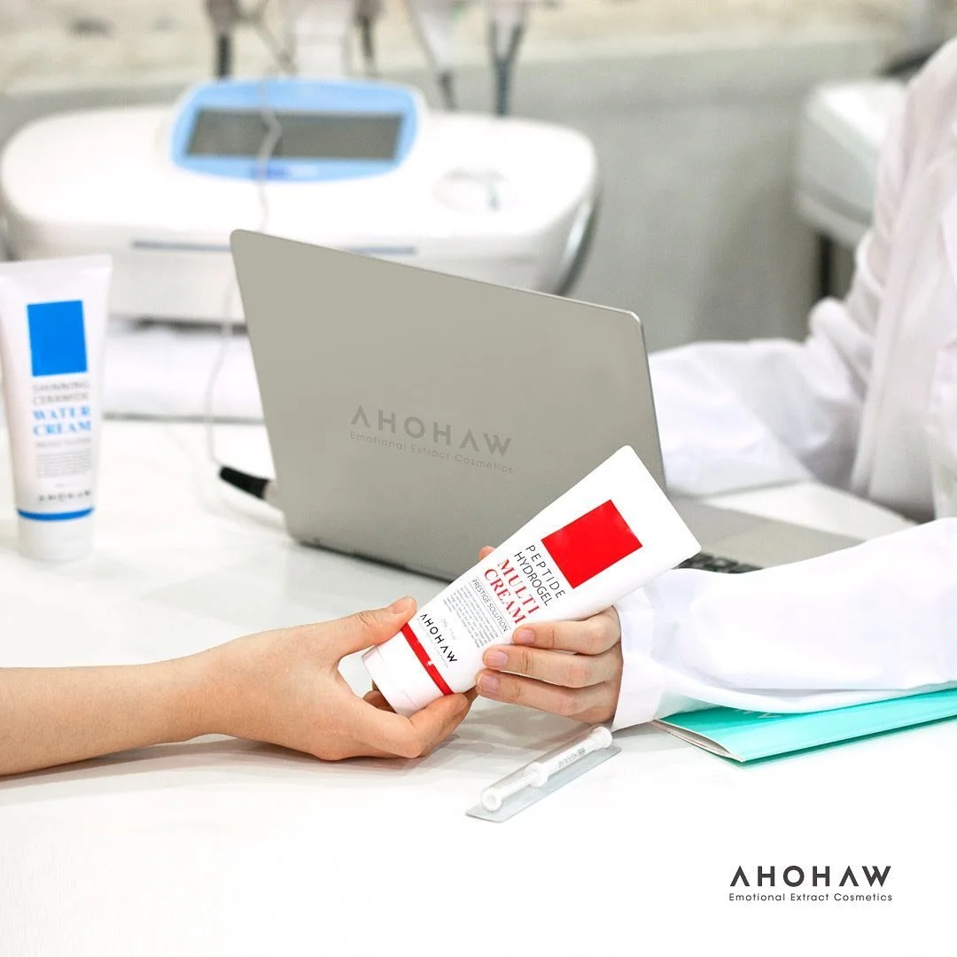 Person handing a tube of AHOHAW cruelty-free moisturizing cream to another person in a clinical setting with a laptop, a tube of hand cream, and a medical device in the background.