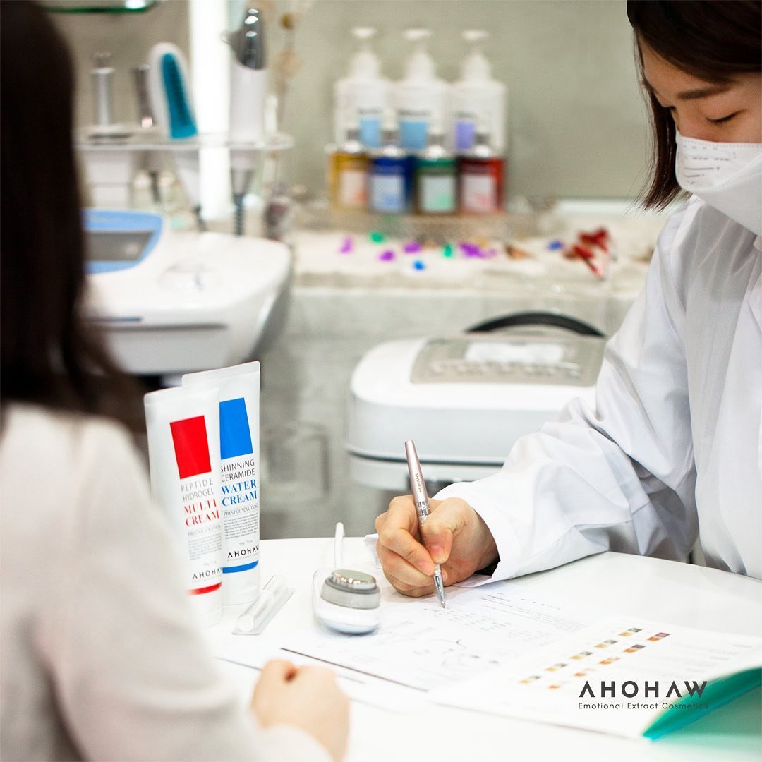A person wearing a face mask and white lab coat writing on paper at a desk with cosmetic products and lab equipment in the background.
