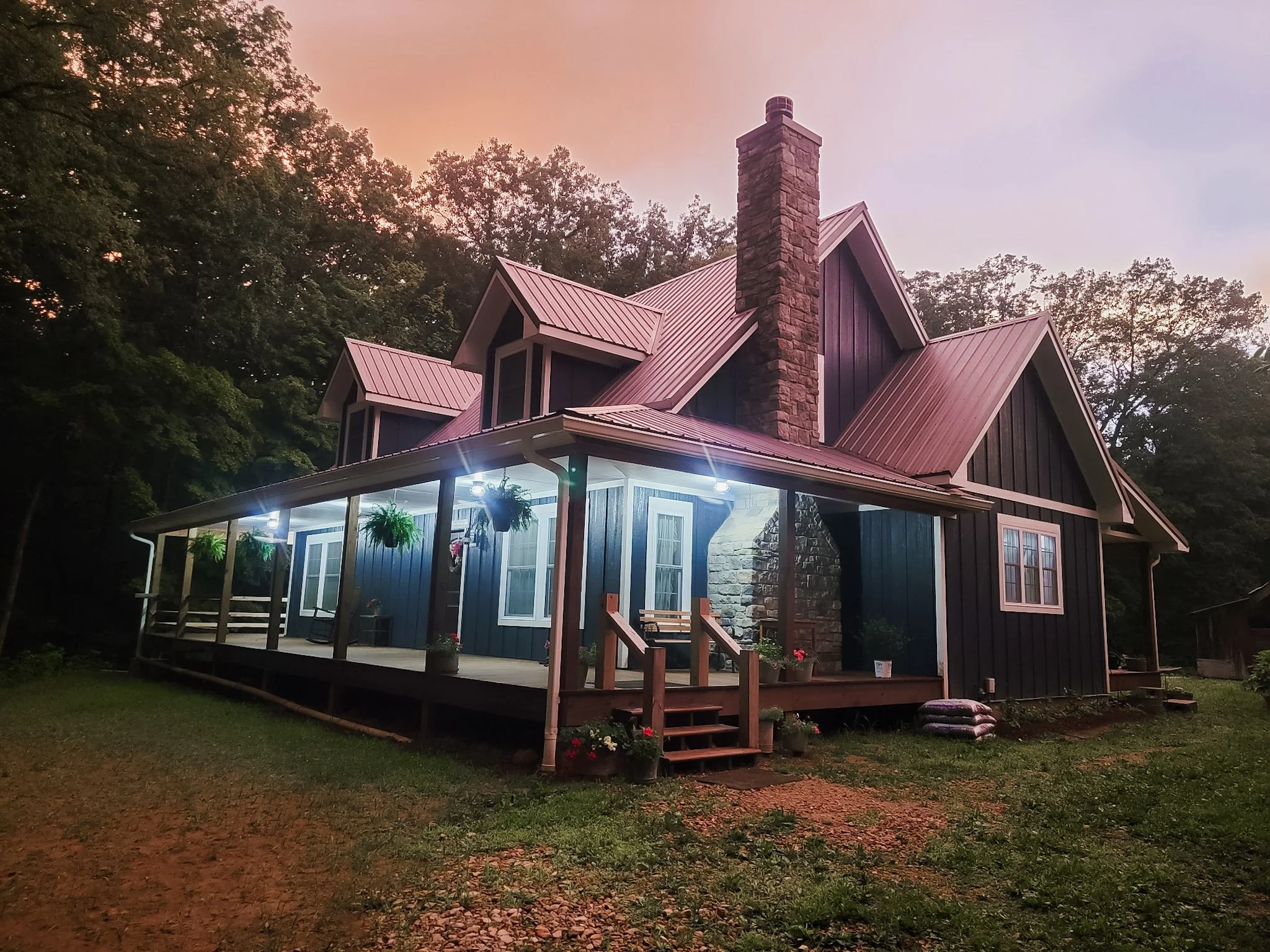 A turnkey, two-story, custom house with a dark blue exterior and a frosted charcol metal roof, featuring a large stone chimney, surrounded by trees at dusk, with a porch illuminated by outdoor lights.