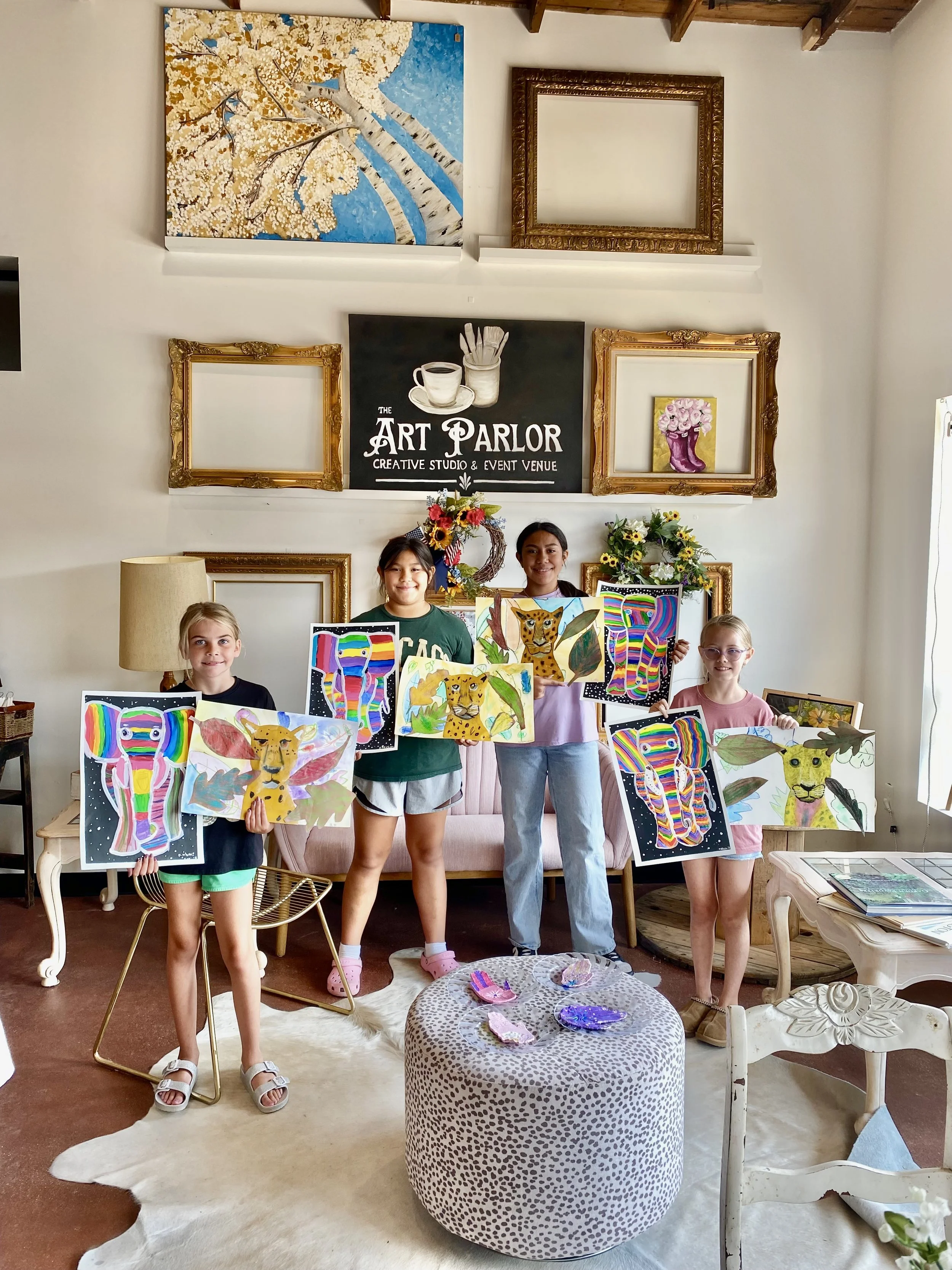 Four young girls stand in an art studio holding colorful paintings of animals, including leopards and butterflies. The studio has artwork and decorative frames on the wall behind them, with a sign reading 'The Art Parlor Creative Studio & Event Venue