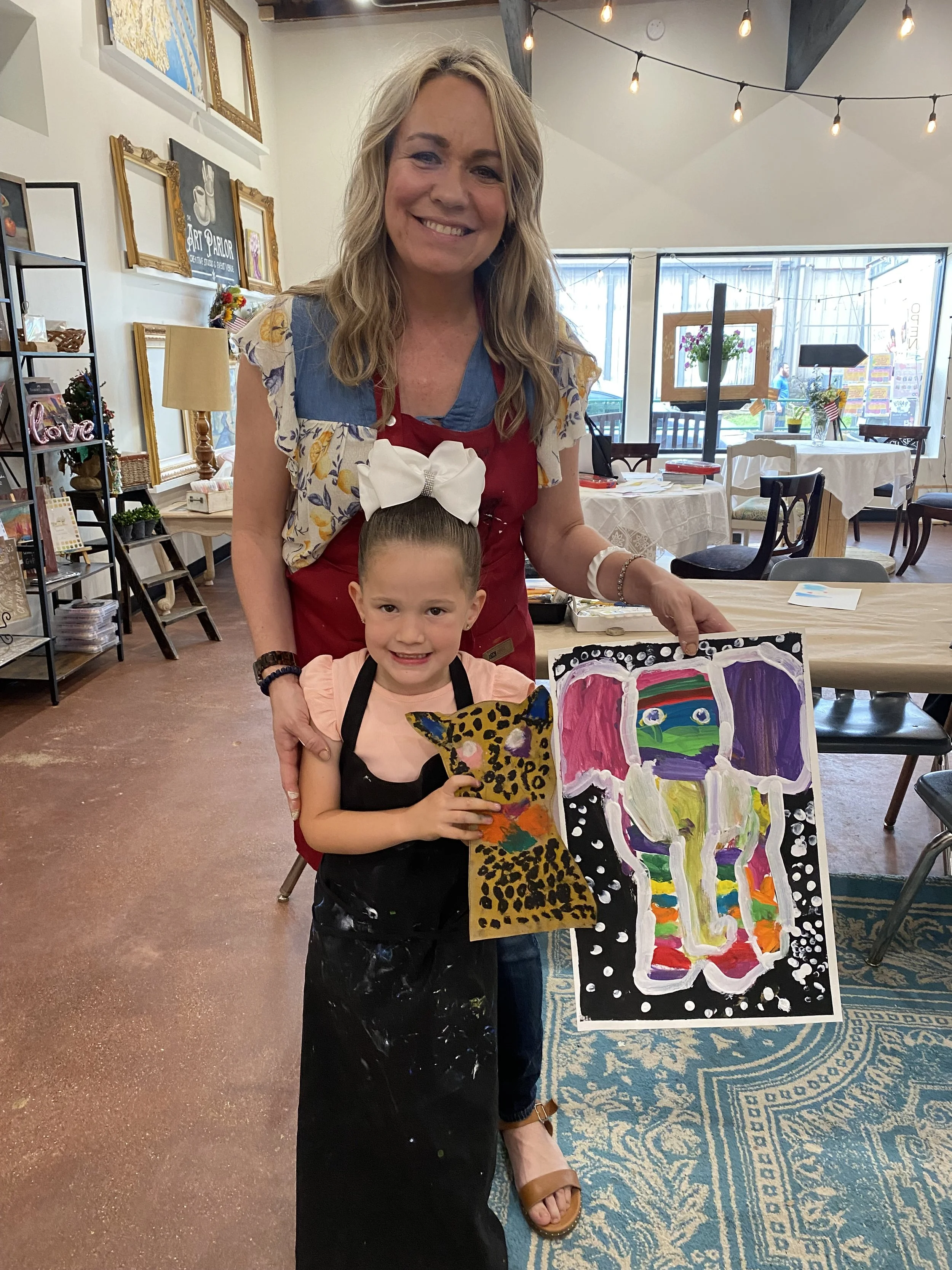 A smiling woman and a young girl holding a colorful painted artwork in a cozy art studio. The girl is holding a painted paper with a leopard pattern and a colorful, abstract artwork resembling a face or figure. The background shows framed artwork, a