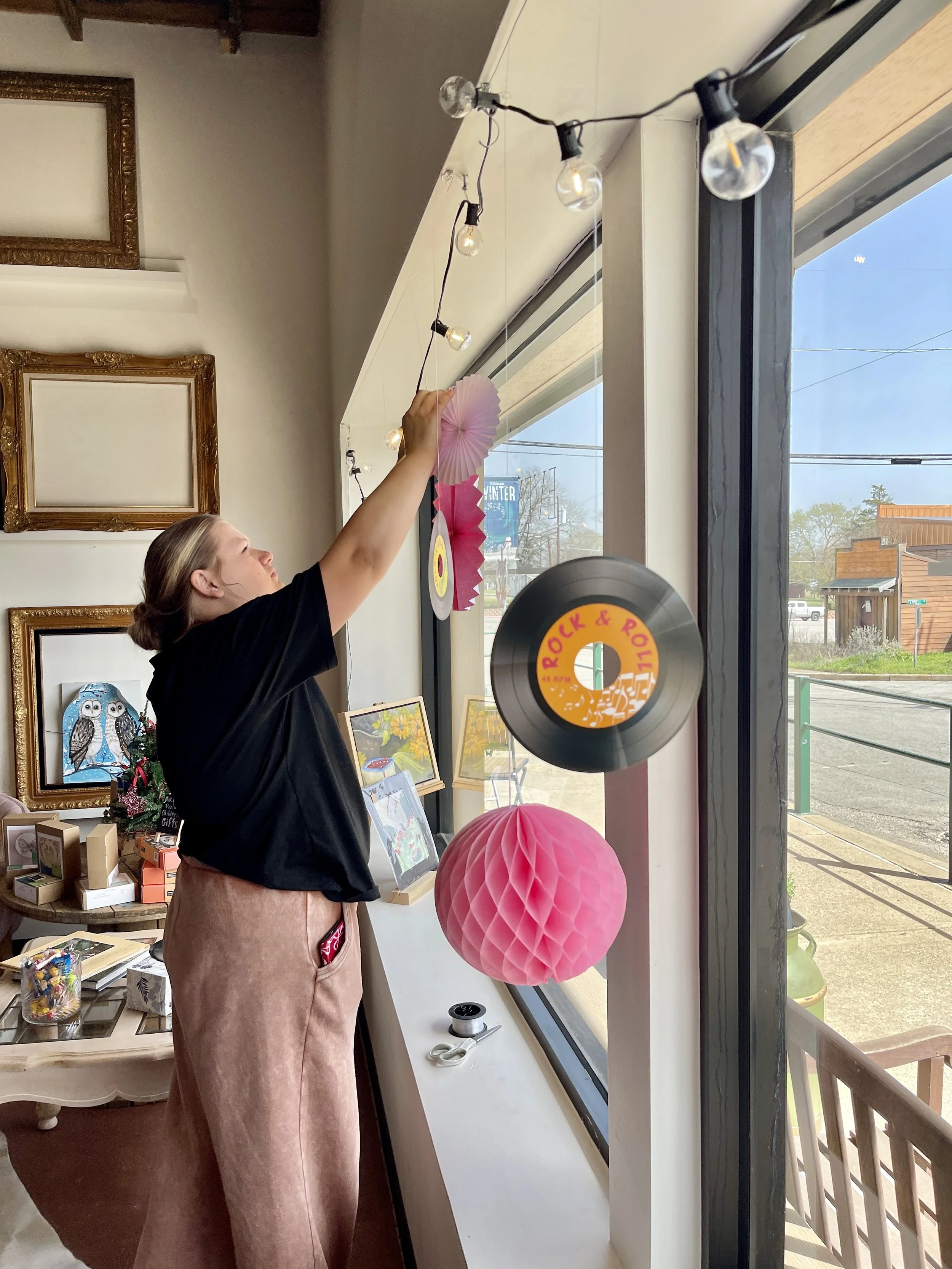A woman decorating a large window with pink paper lanterns, hanging string lights, and colorful paper decorations in a room with framed artwork and a table with various craft supplies.