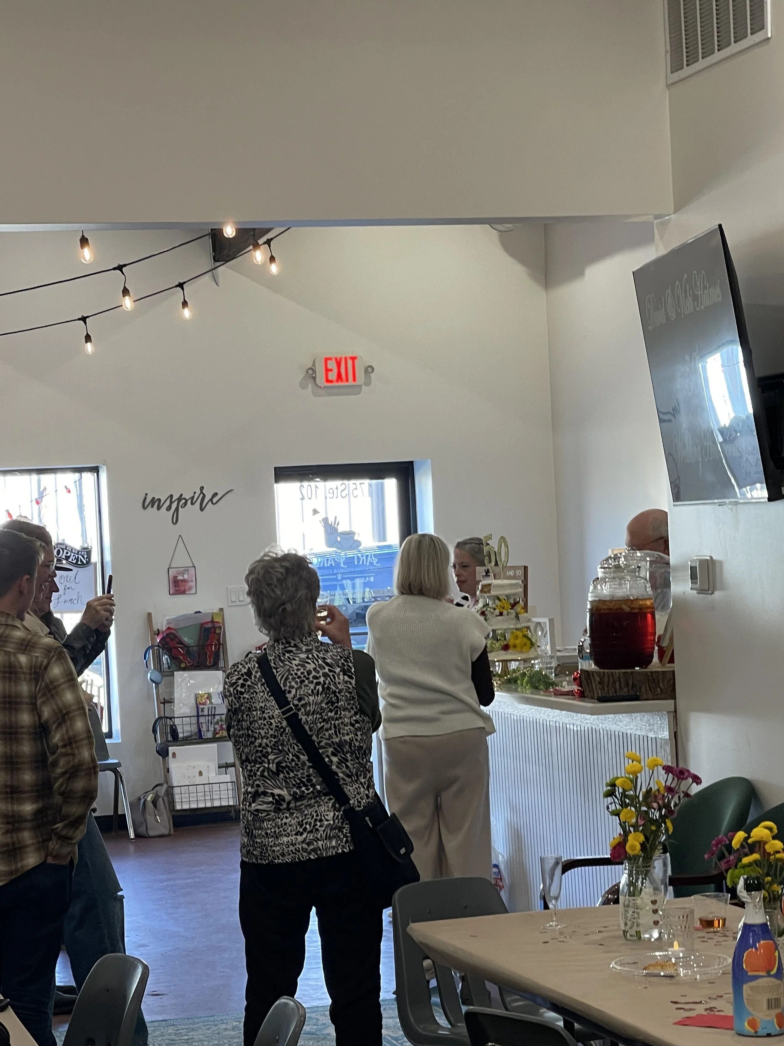 People standing in line or waiting at a counter inside a cafe or bakery with a sign that says 'inspire' on the wall, colorful flowers on a table, and string lights hanging from the ceiling.
